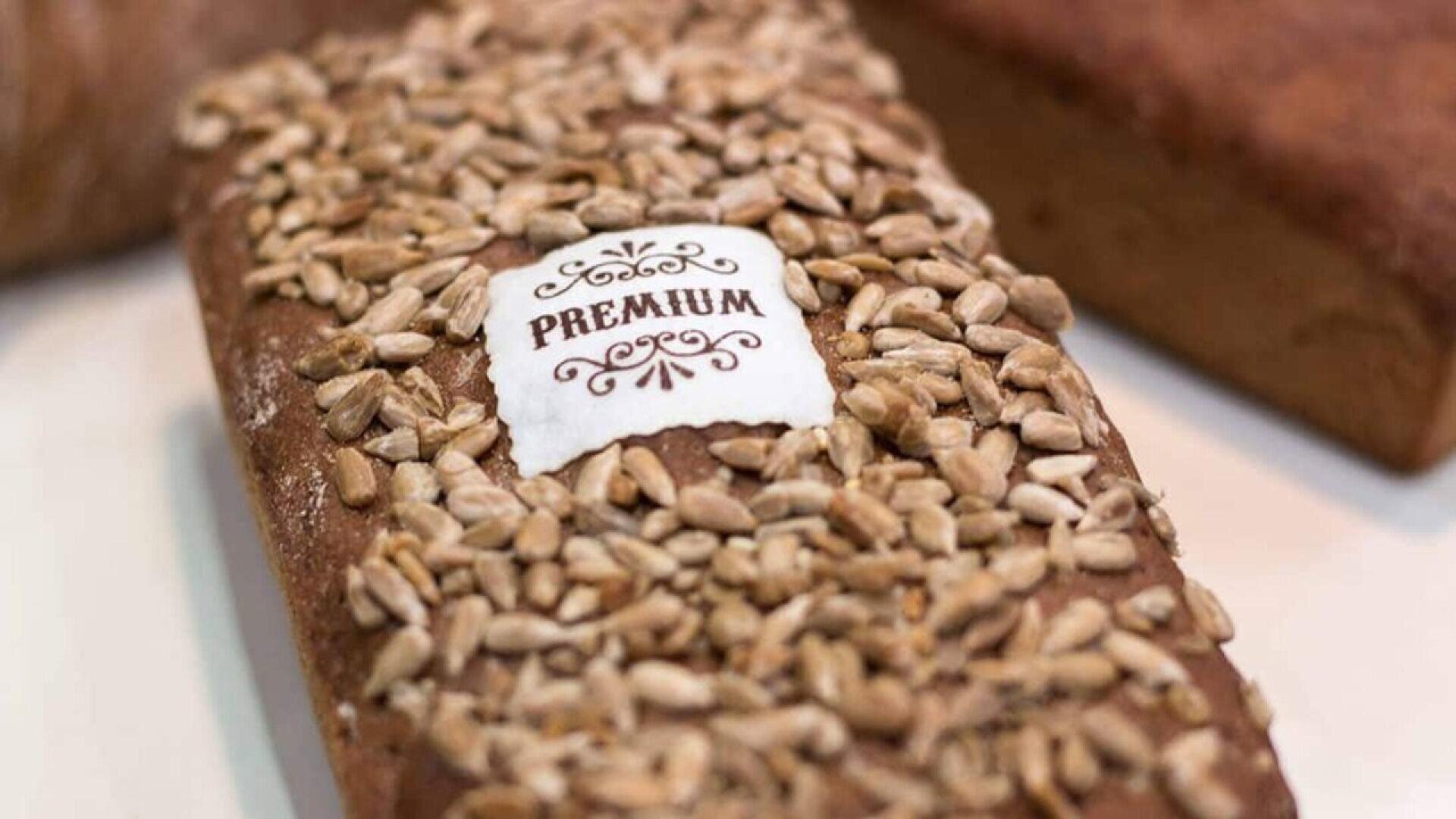 Ein Laib Brot mit Sonnenblumenkernen und einem weißen Etikett mit der Aufschrift PREMIUM in dekorativer Schrift. Das Brot liegt auf einer hellen Fläche, im Hintergrund ist ein Teil eines anderen Brotes zu sehen.