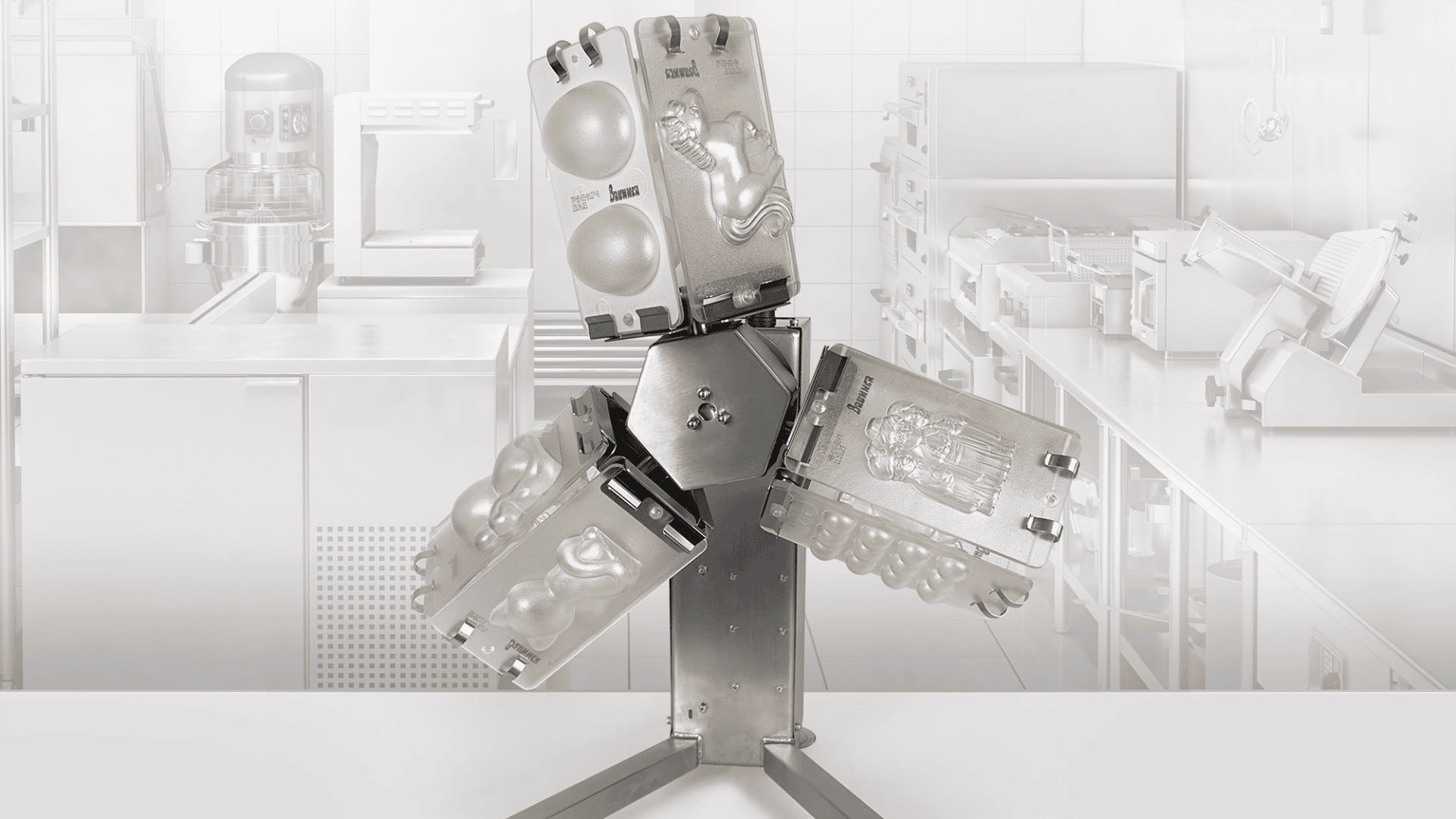 A commercial kitchen appliance with four metal molds attached, each shaped for making different 3D chocolate figures, positioned on a metal stand with a kitchen background.