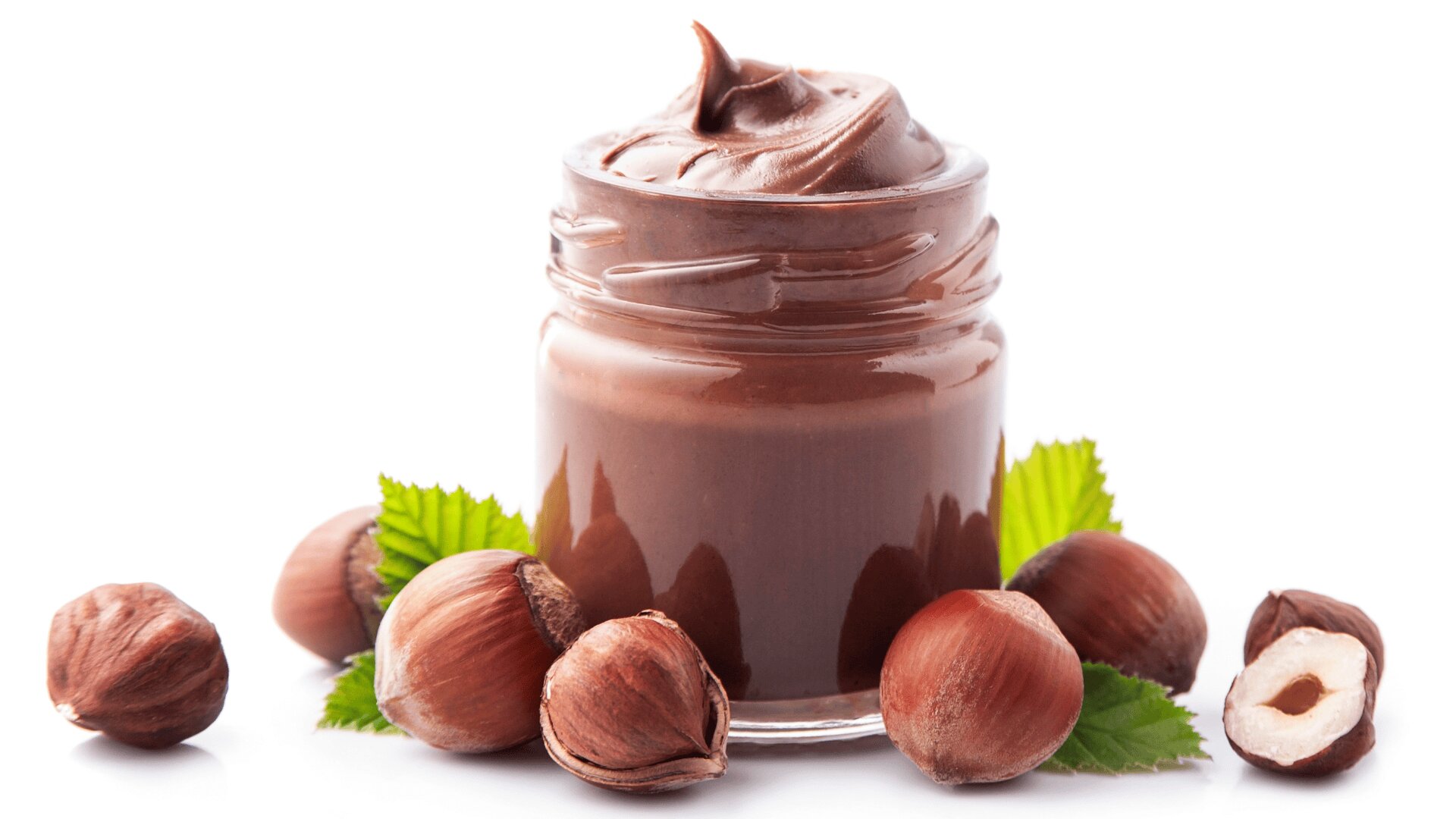 A glass jar filled with creamy chocolate hazelnut spread, surrounded by whole and cracked-open hazelnuts and green leaves, all set against a white background.