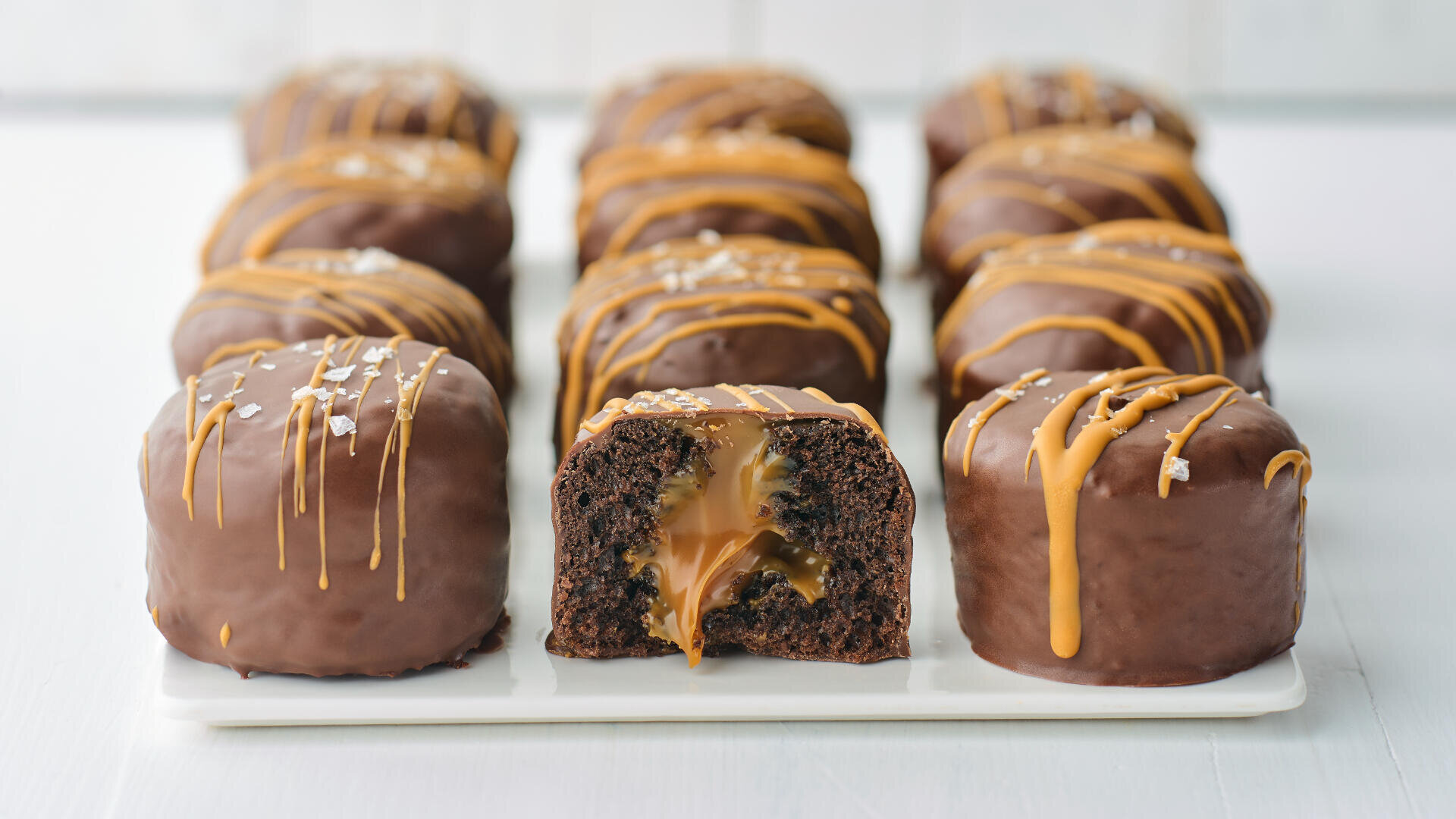 Rows of round chocolate cakes topped with caramel drizzle and sea salt are displayed on a white tray. The center cake is cut open, revealing gooey caramel filling inside the rich chocolate cake.