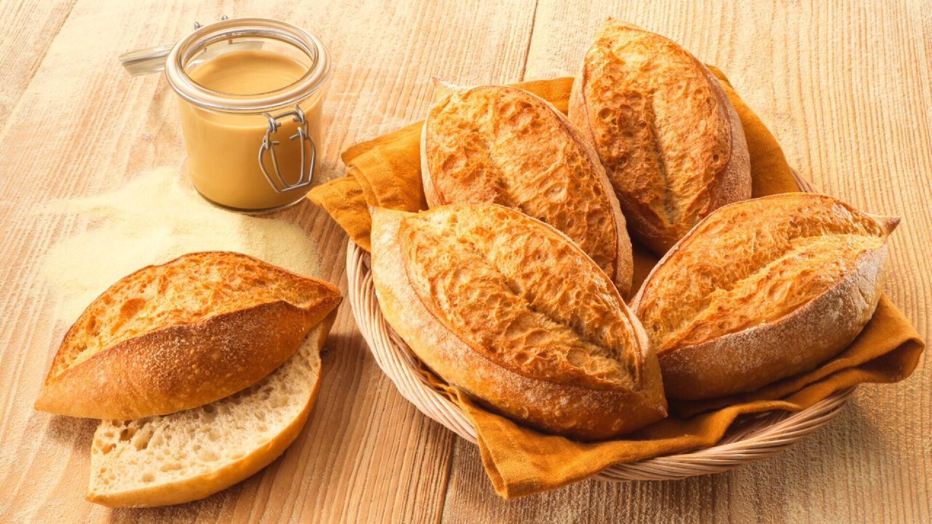 A wicker basket lined with a yellow cloth holds five crusty bread rolls. One roll is sliced in half on the table next to the basket. A jar of creamy spread sits nearby on a wooden surface.