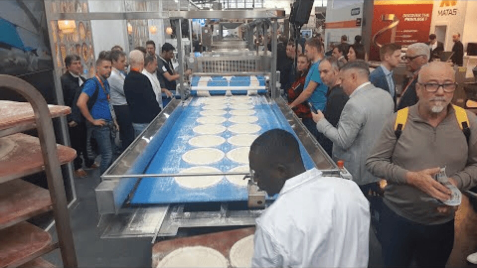 A group of people watch a factory line where circles of pizza dough move along a conveyor belt; some workers handle the dough while others watch in a busy hall.