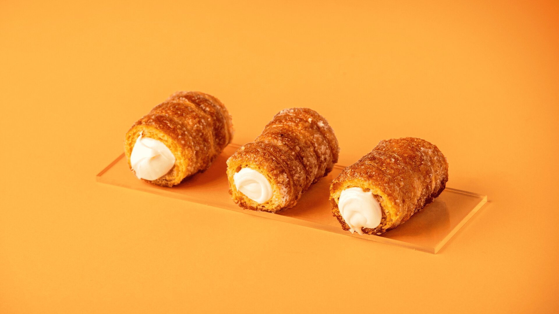 Three cream-filled pastry rolls coated with sugar are arranged in a row on a transparent rectangular tray, set against an orange background.
