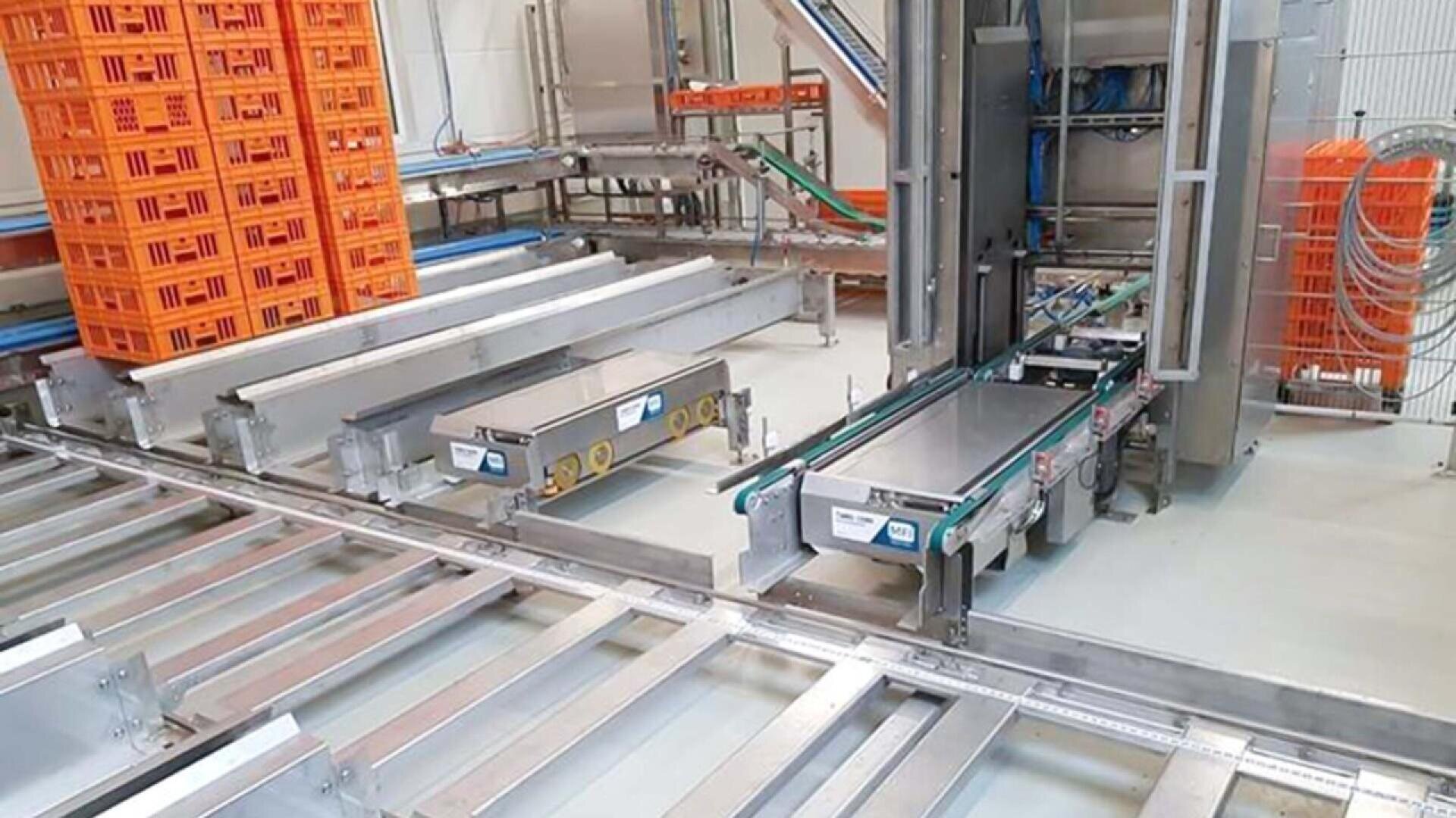 Automated conveyor system in a factory with empty metal tracks and orange plastic crates stacked in the background, designed for transporting goods or materials.