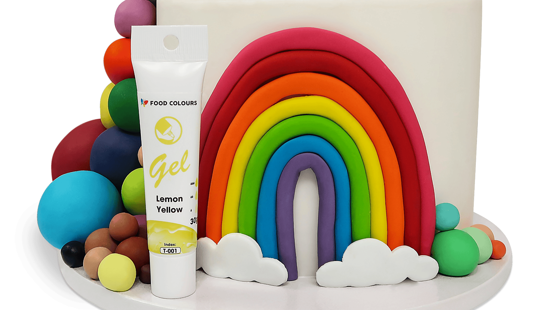 A white cake decorated with a colorful rainbow and clouds made of fondant, surrounded by colorful fondant balls, with a tube of lemon yellow gel food coloring placed in front.