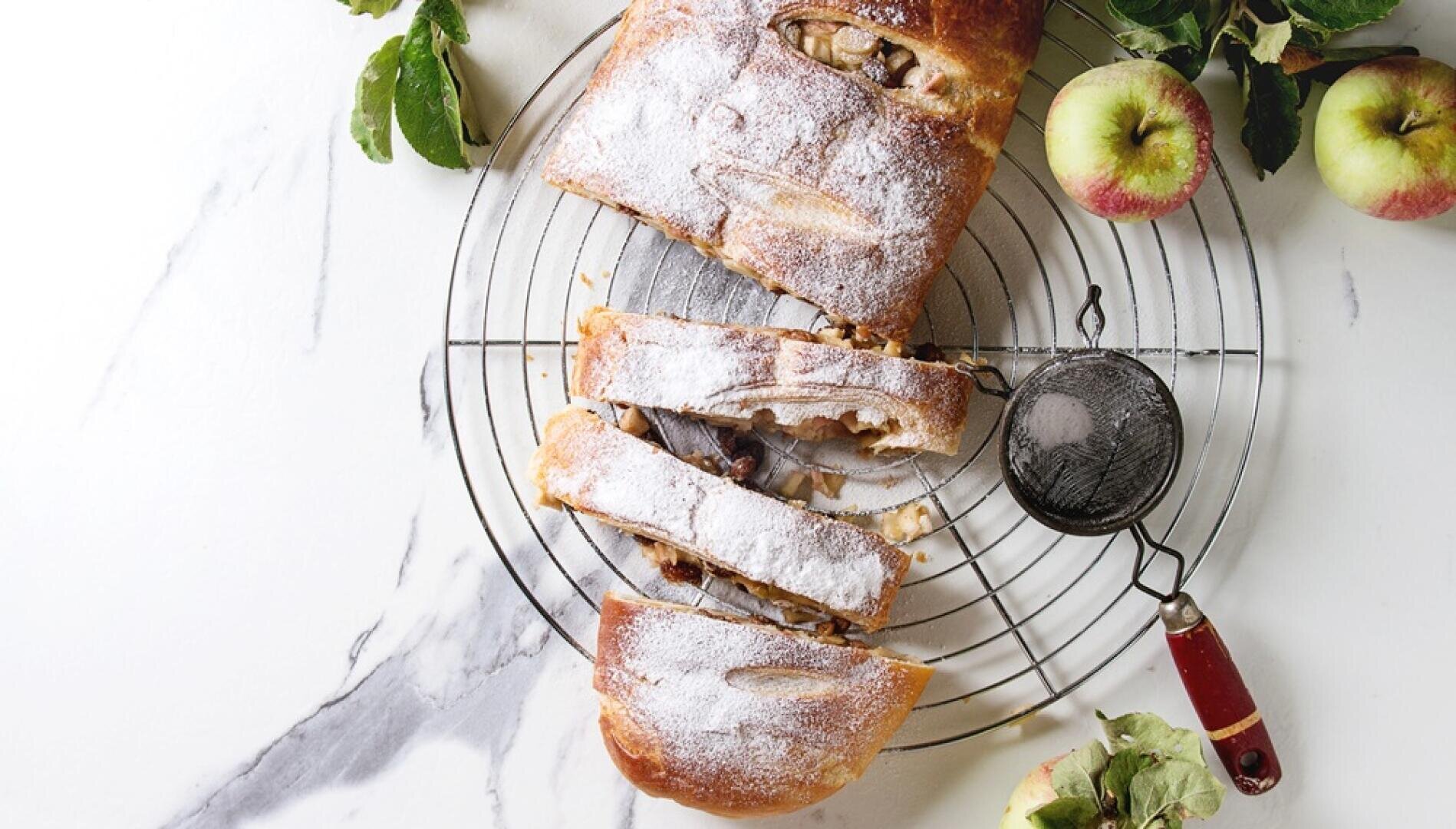 Ein in Scheiben geschnittener, mit Puderzucker bestäubter Apfelstrudel liegt auf einem Kuchengitter. Daneben stehen auf einer weißen Marmorplatte ein Sieb mit Zucker, grünen Äpfeln und grünen Blättern.