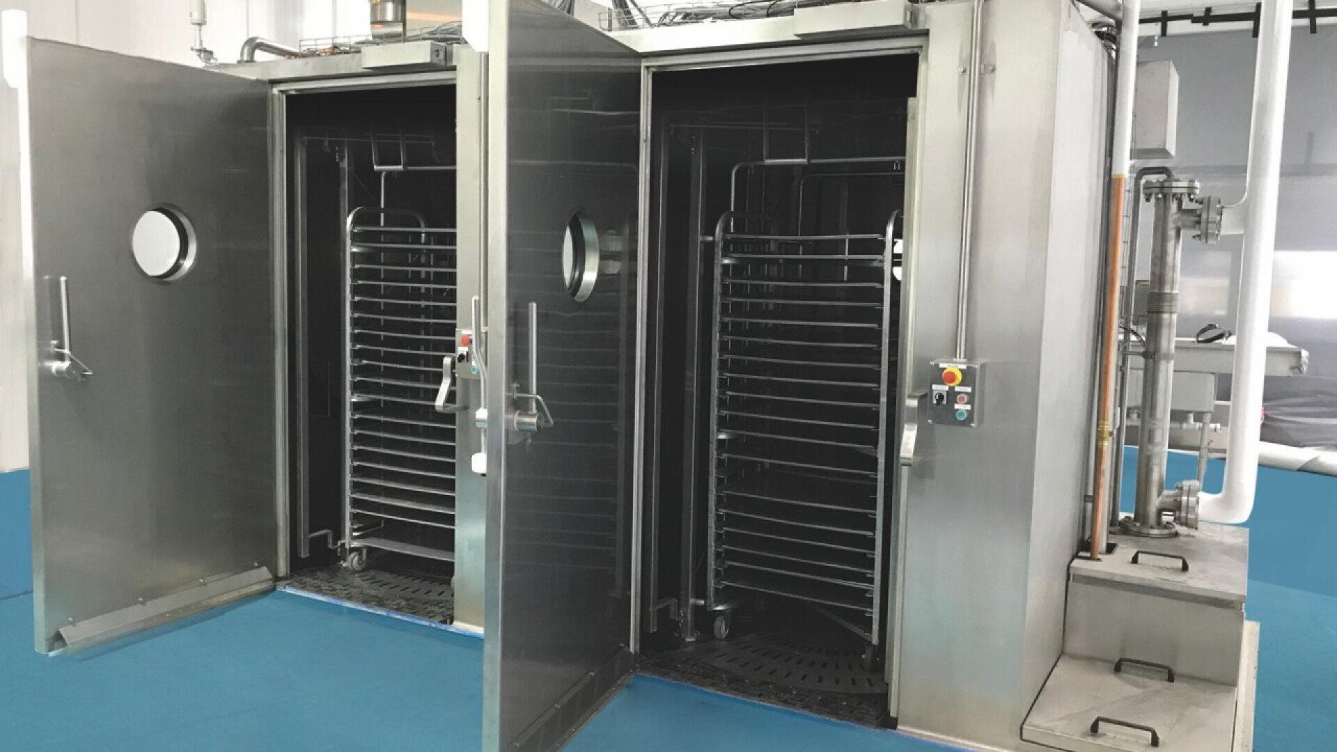 Two large, stainless steel industrial ovens with open doors reveal empty interior racks. The ovens are set on a blue floor in a clean, modern facility with control panels and pipes visible on the right side.