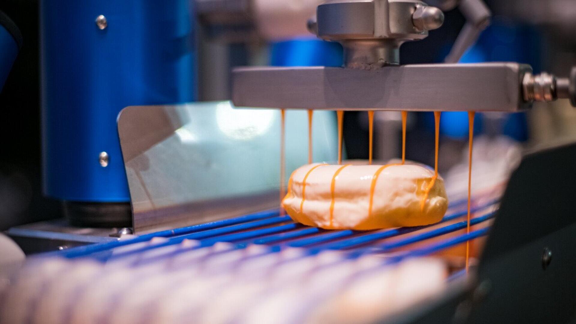 A donut is coated with icing as it moves along a conveyor belt in a factory, with orange icing being drizzled from a machine onto the pastry.