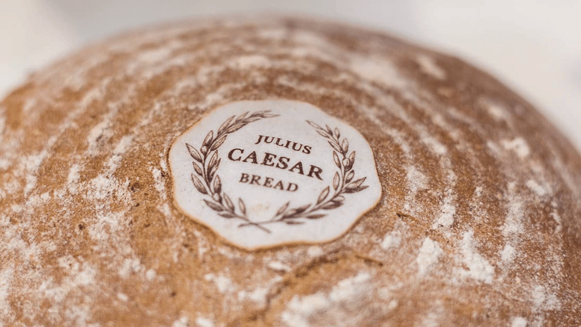 A round loaf of bread with a circular white label on top that reads Julius Caesar Bread surrounded by a laurel wreath design. The bread has a dusting of flour and a rustic, textured crust.