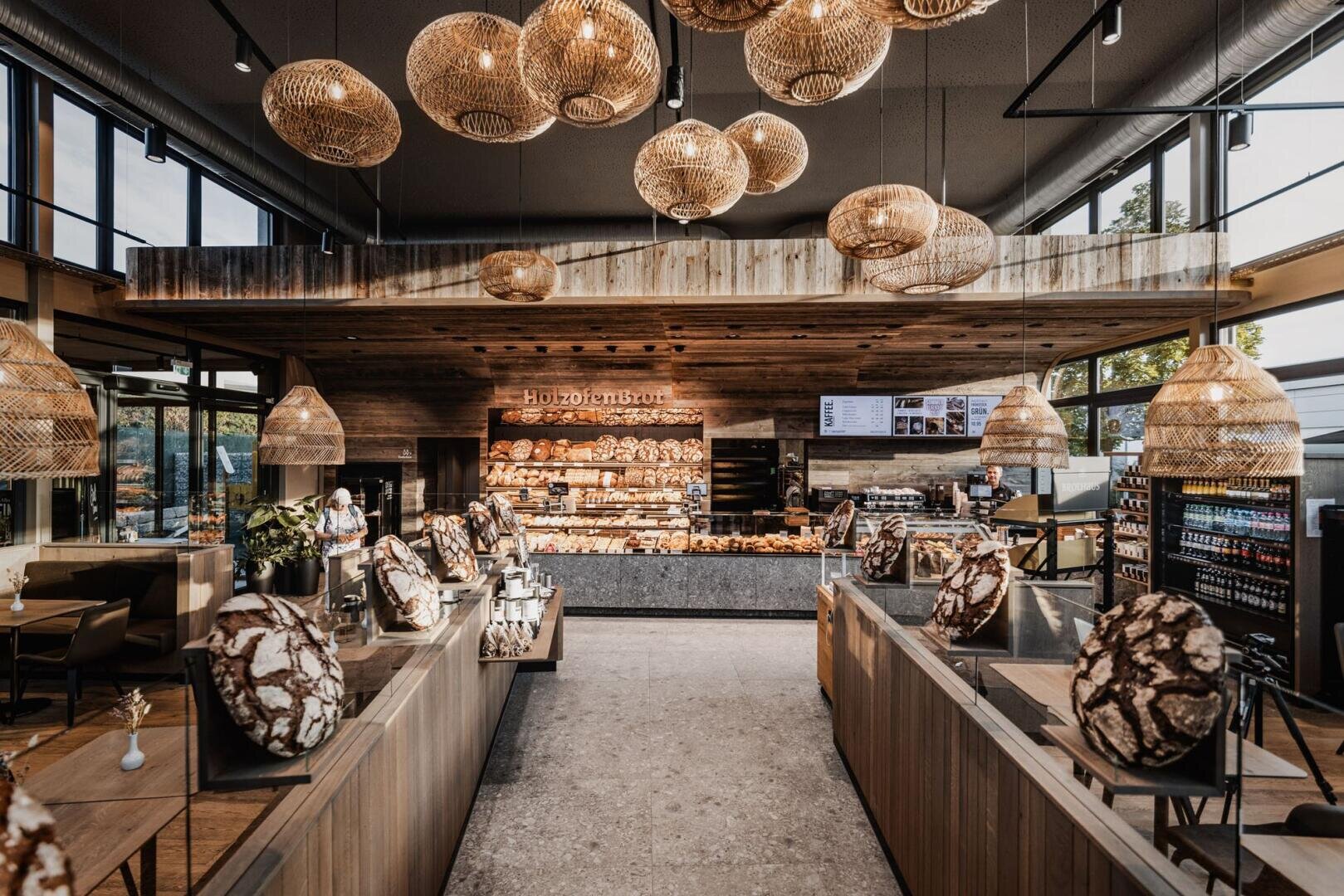 Eine moderne Bäckerei mit Holzakzenten, großen Korblampen und rustikalen, runden Brotlaiben. Hinter der Theke sind Regale mit verschiedenen Backwaren gefüllt. Große Fenster sorgen für Tageslicht.