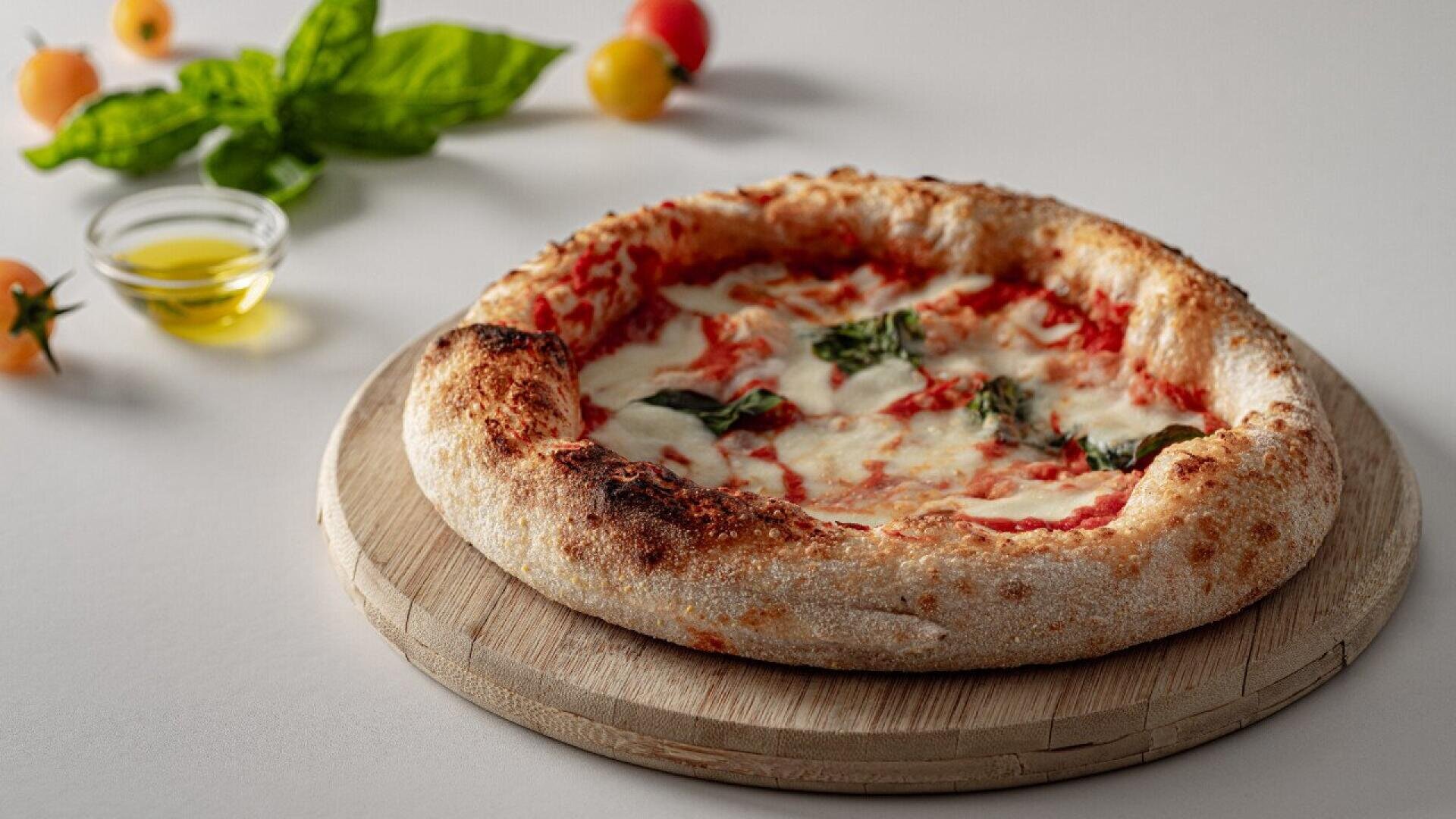 A Neapolitan-style Margherita pizza with melted cheese, tomato sauce, and basil sits on a wooden board. Nearby, there are fresh basil leaves, cherry tomatoes, and a small bowl of olive oil on a light surface.
