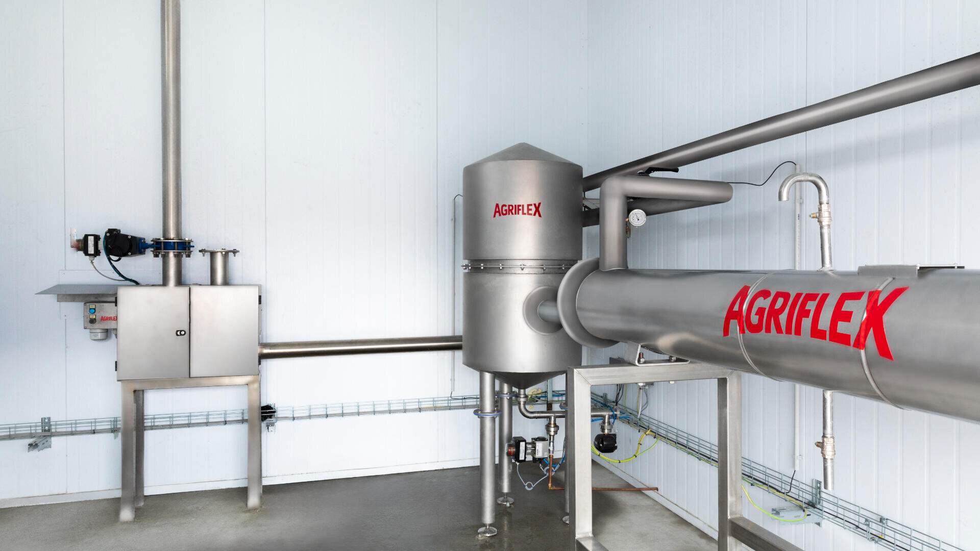 A stainless steel industrial processing system labeled AGRIFLEX, with pipes and cylindrical tanks, is installed in a clean, white-walled room with visible control panels and wiring on the wall.