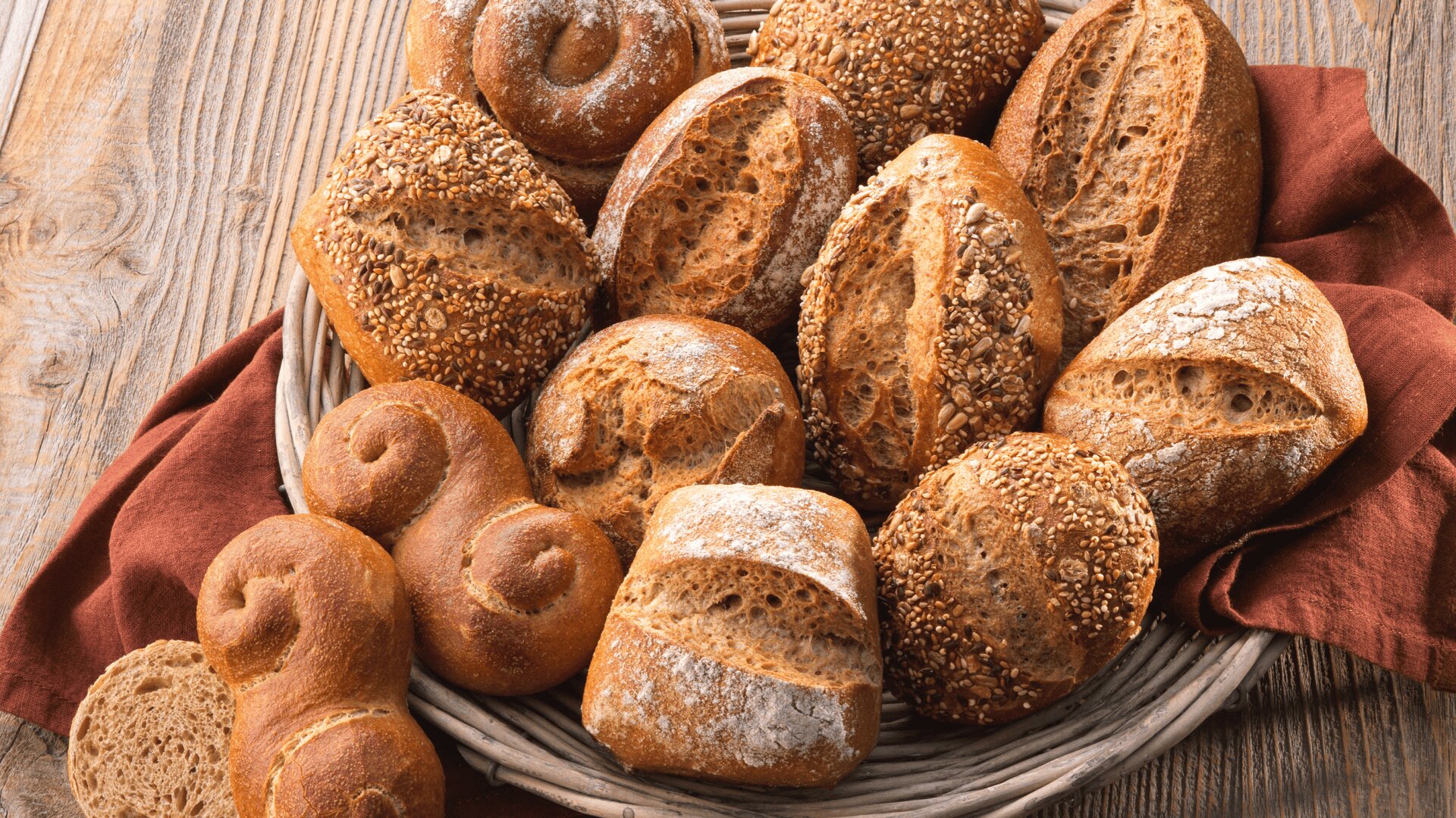 Ein Weidenkorb mit verschiedenen rustikalen Brötchen, manche mit Körnern, manche mit Mehl bestäubt, steht auf einem roten Tuch auf einem Holztisch. Zwei geschnittene Brötchen und ein dekoratives Schneckenbrötchen stehen davor.