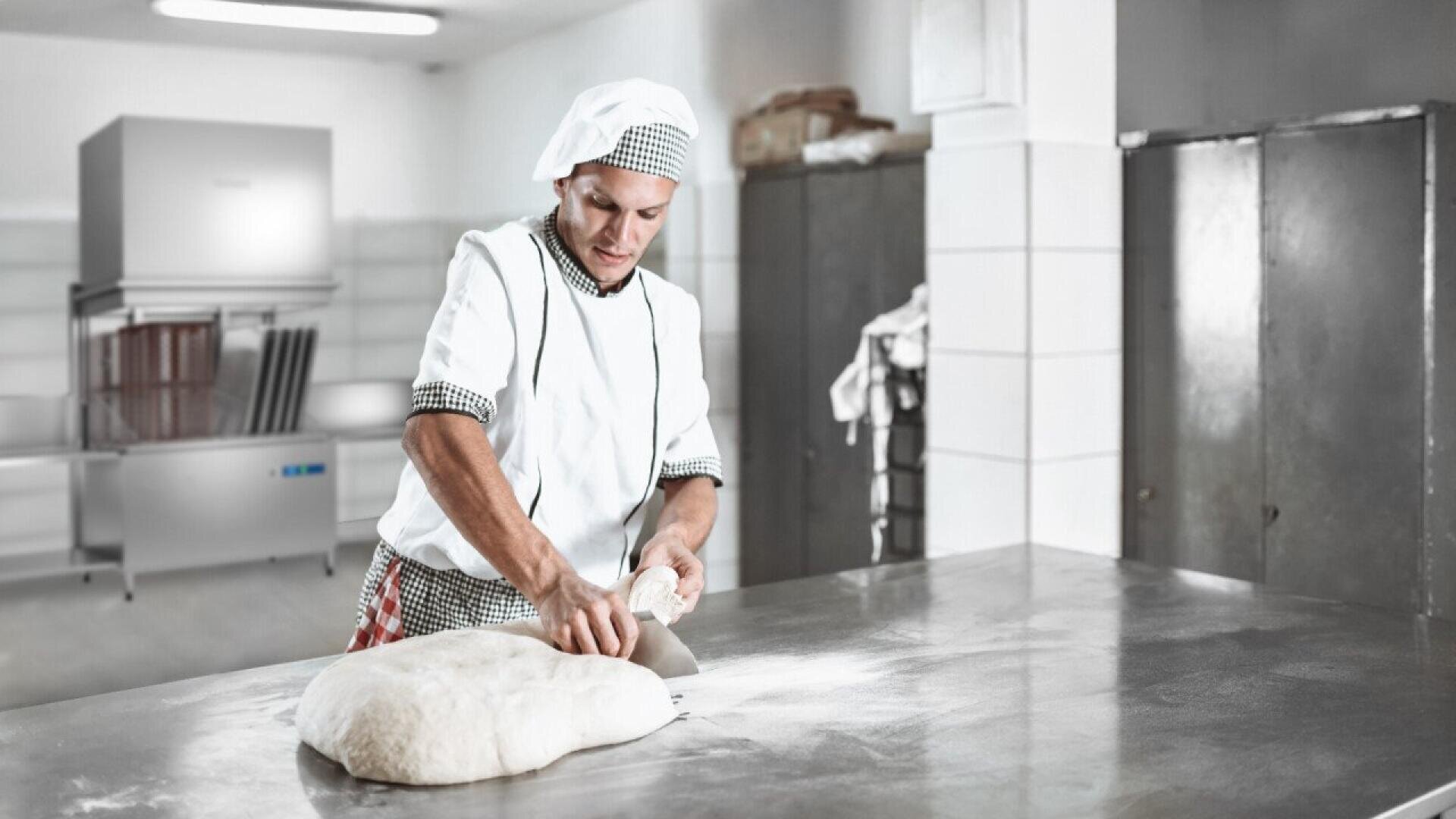 A baker wearing a white uniform and hat kneads dough on a floured stainless steel countertop in a commercial kitchen.