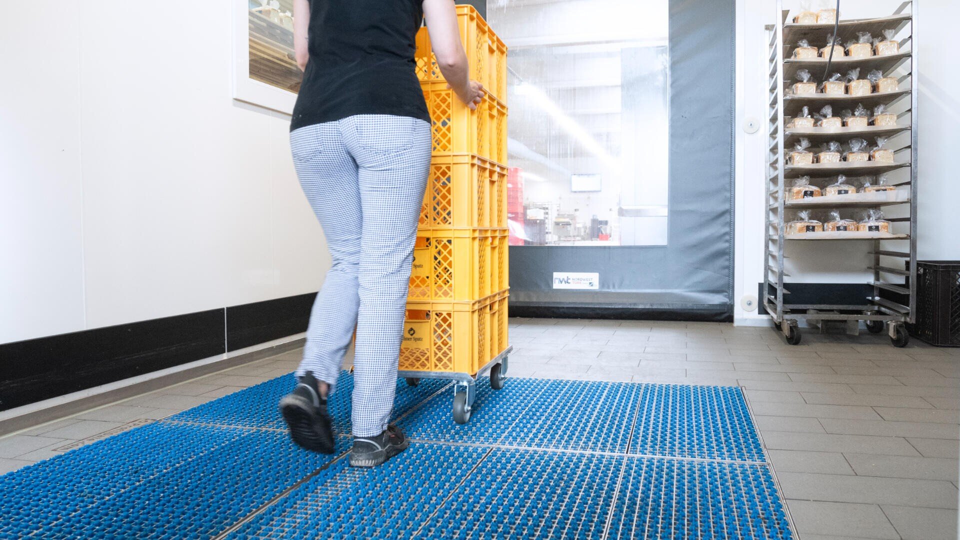 Person pushing a cart stacked with yellow crates across a blue anti-slip mat in a clean, industrial setting; shelves with small items are visible in the background.