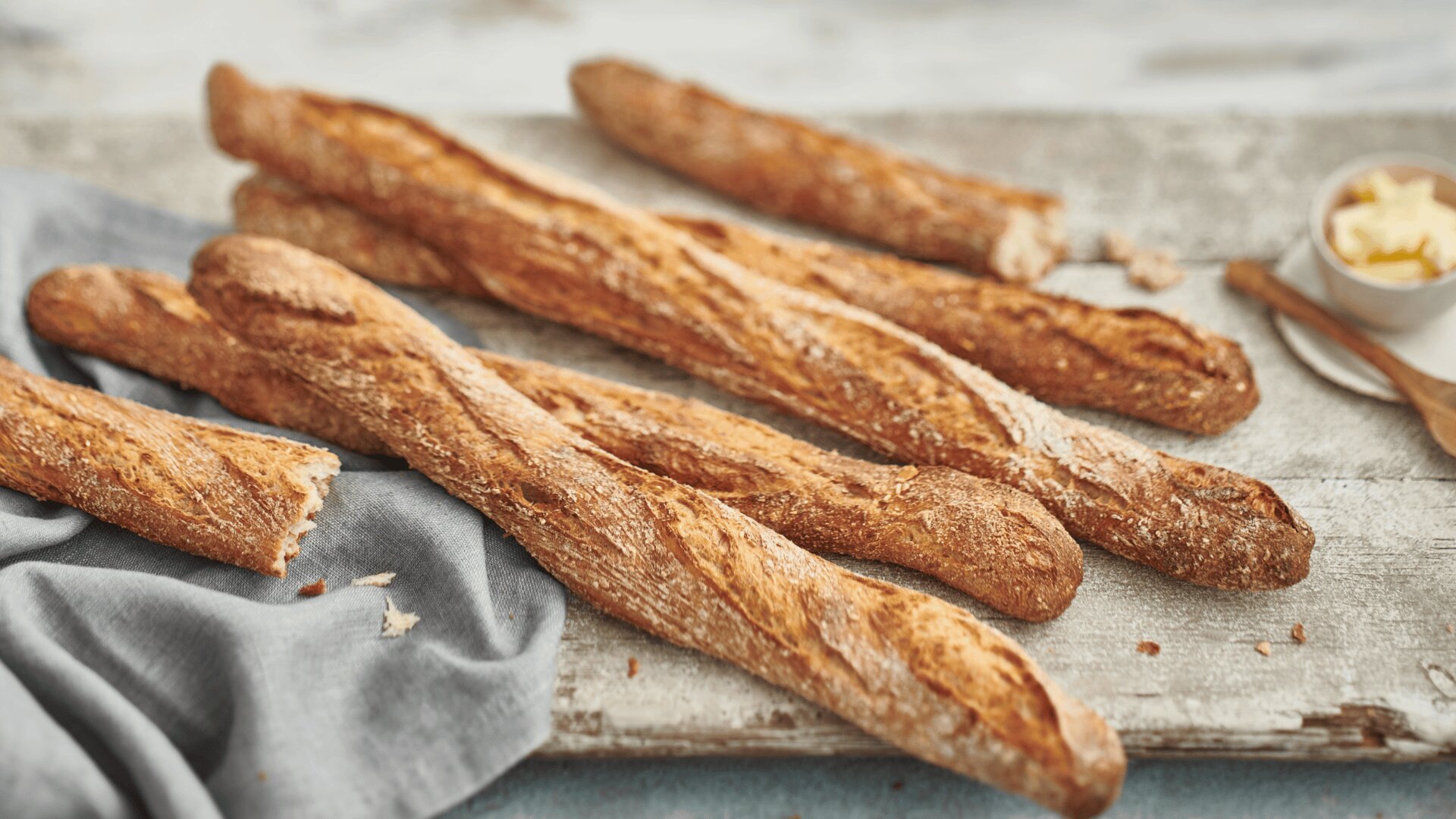 Mehrere frisch gebackene Baguettes liegen auf einer rustikalen Holzplatte neben einem grauen Tuch, im Hintergrund eine kleine Schale mit Butter und ein Holzmesser.