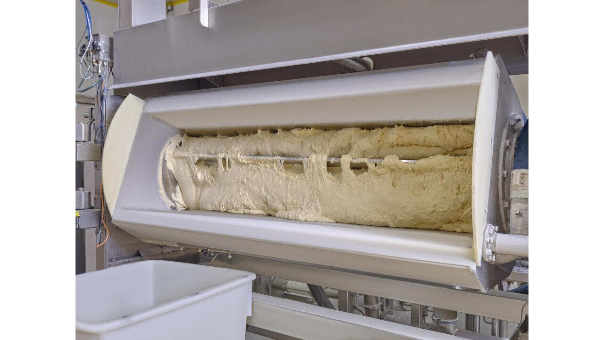 Ein großer industrieller Teigkneter mit einer rotierenden Trommel wird mit Rohteig gefüllt. Der Kneter ist aus rostfreiem Stahl und befindet sich über einem weißen Behälter zum Auffangen des gemischten Teigs.