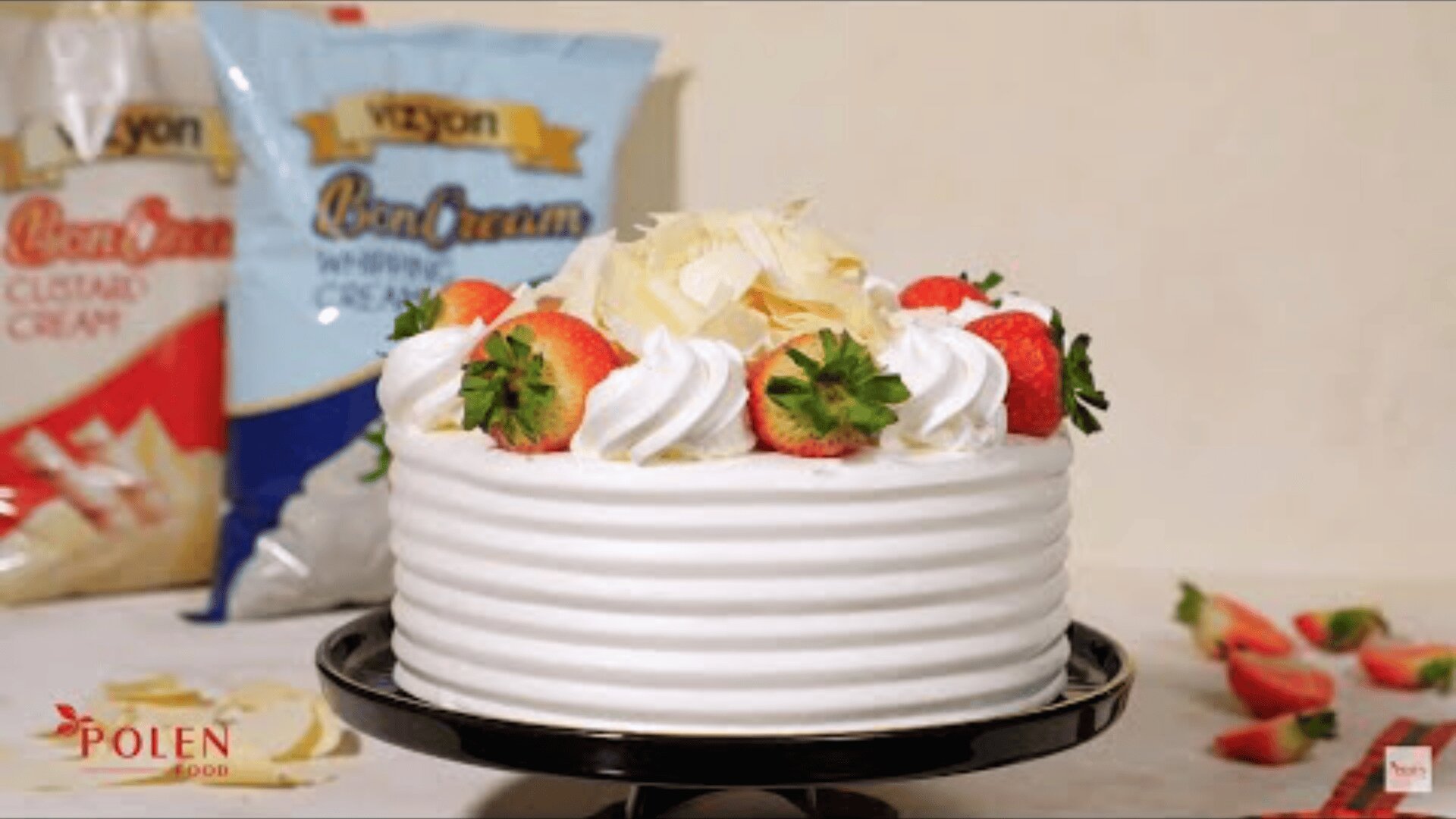 A white frosted cake topped with whipped cream, strawberries, and white chocolate curls sits on a black cake stand. There are packets of cream in the background and strawberry slices nearby.