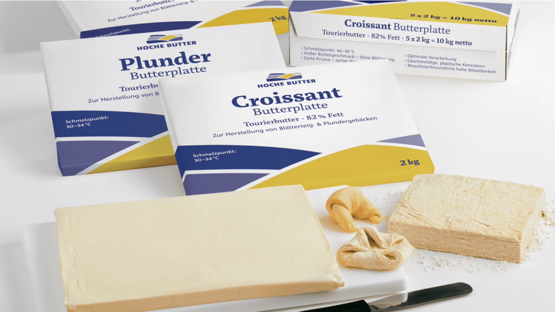 Several packages of German baking butter labeled Croissant Butterplatte and Plunder Butterplatte are shown beside a slab of butter, a block of laminated dough, and two rolled croissant pastries on a white surface.