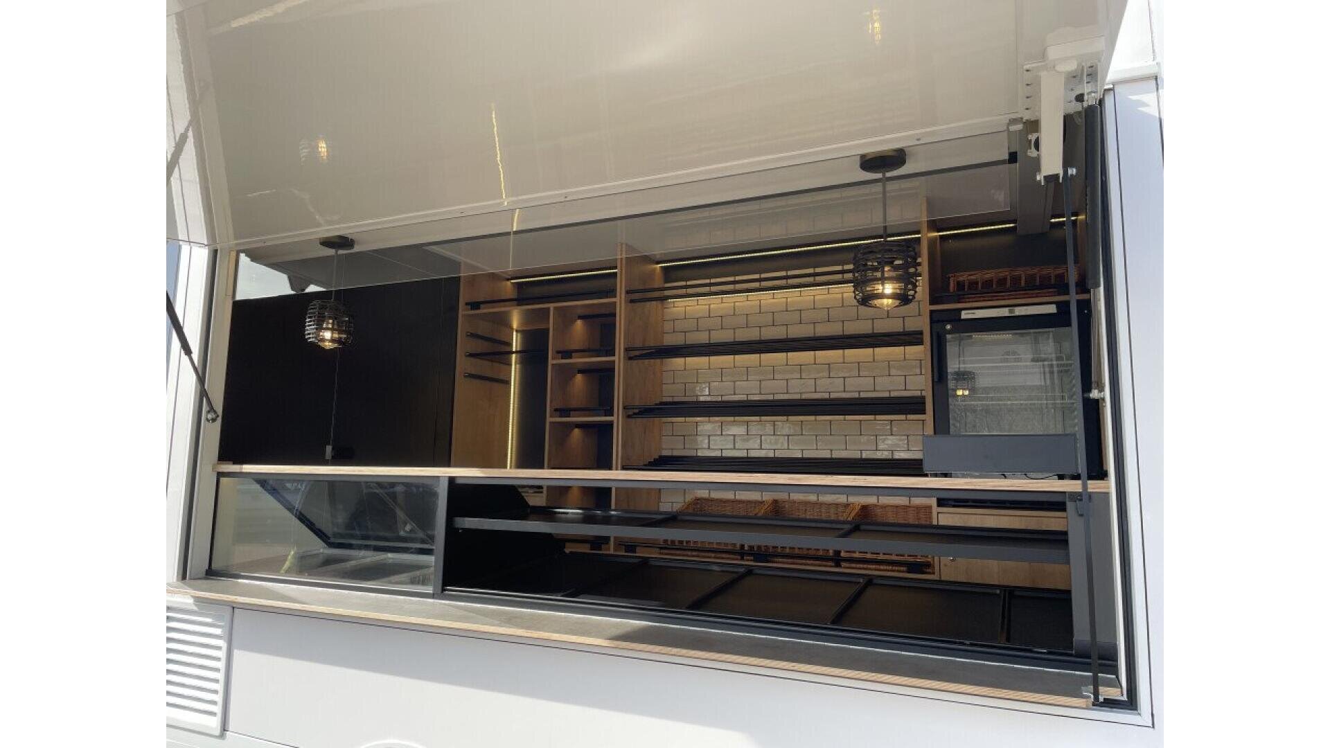 The image shows the open serving window of a modern food truck or trailer with empty shelves, wicker baskets, white subway tile backsplash, and minimalistic lighting. No food or people are visible inside.