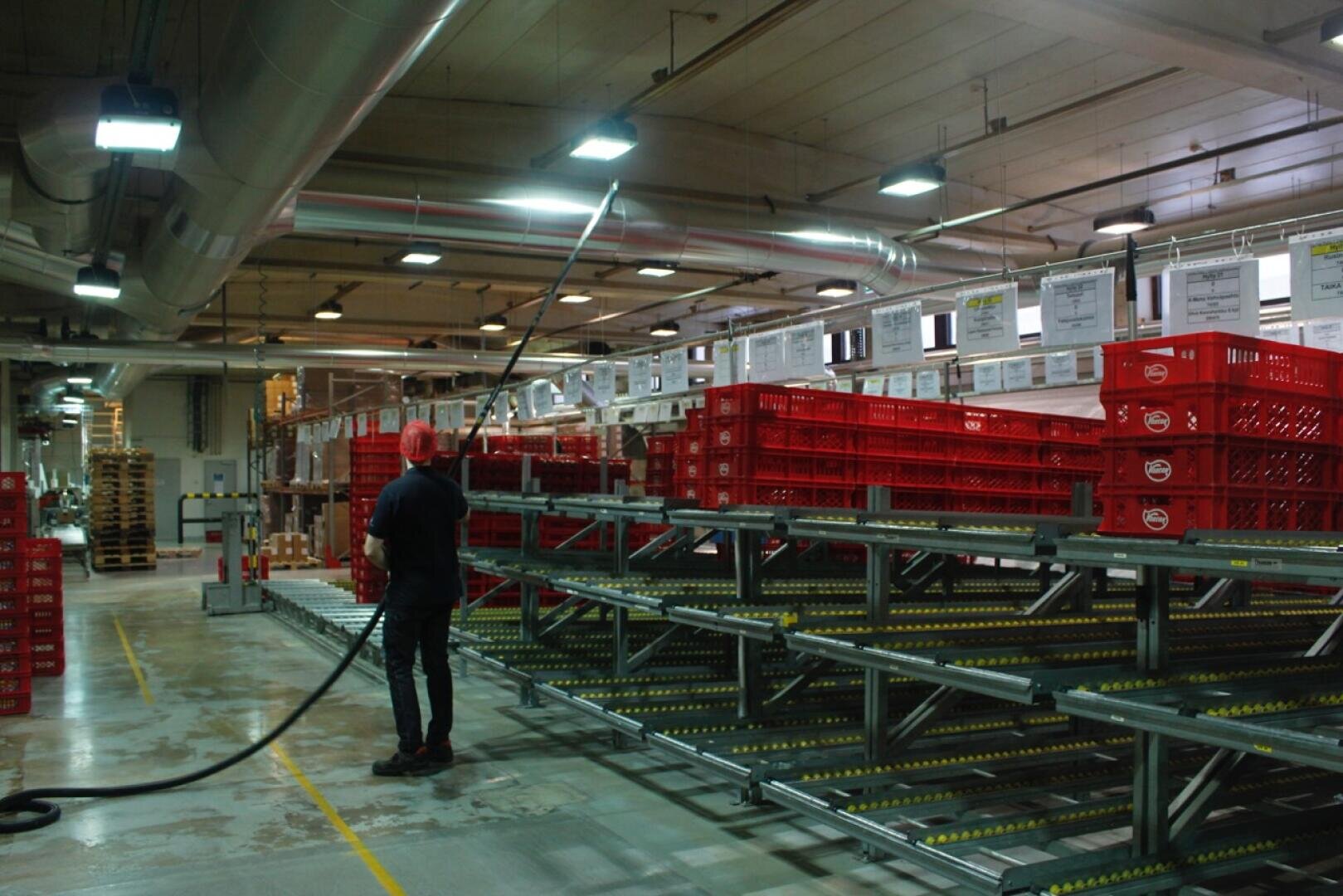 A person in a warehouse stands near empty roller shelves and red crates, holding a long hose or cable. The warehouse has overhead lighting, industrial equipment, and informational signs above shelves.