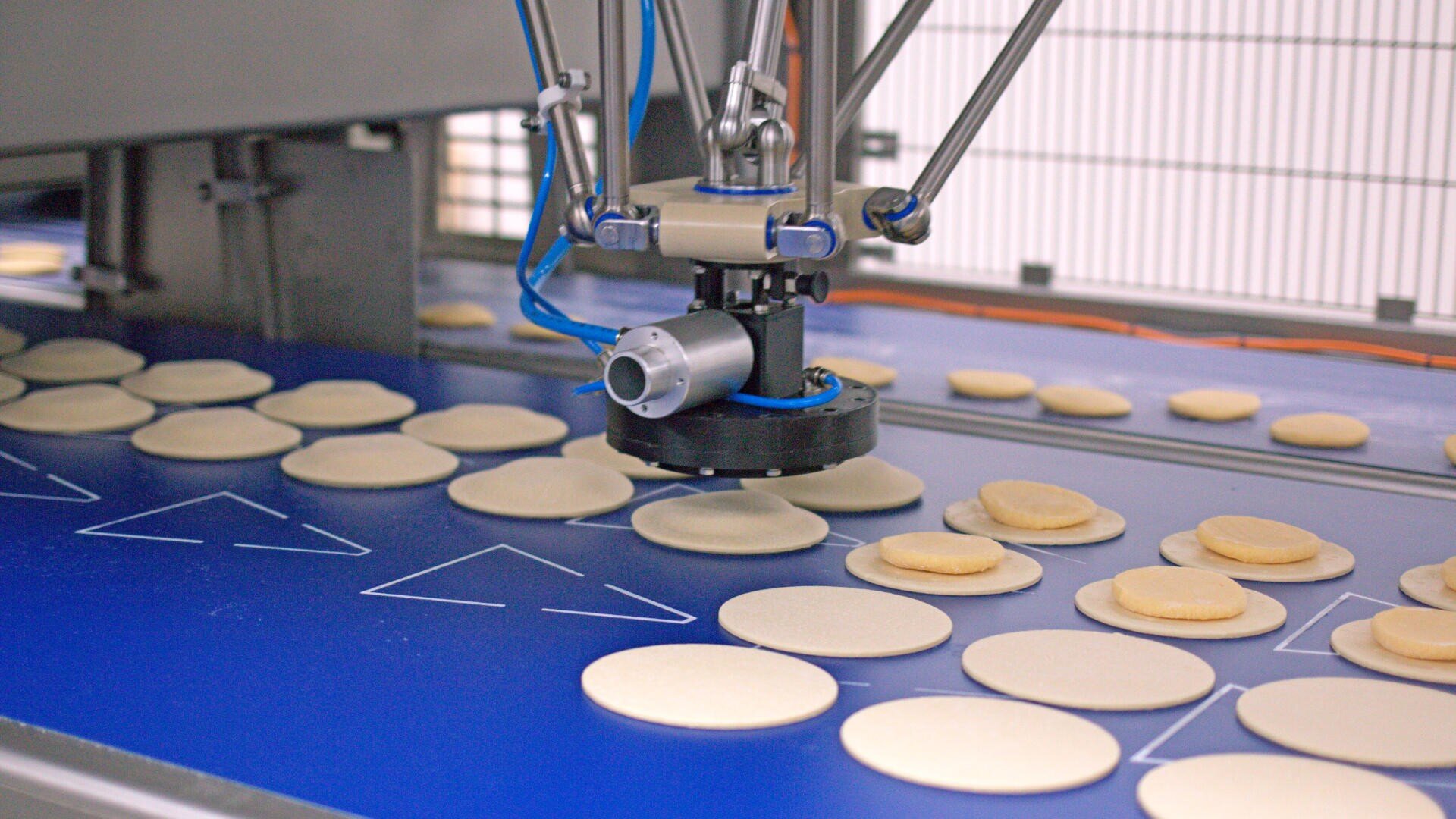 A robotic arm places dough rounds on a conveyor belt in a food processing factory, assembling stacks of dough pieces for further production.