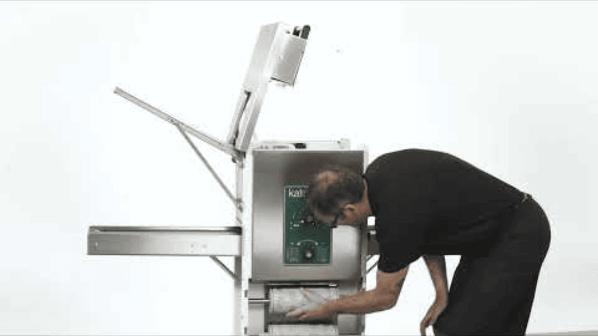 Eine Person in schwarzer Kleidung beugt sich zum Be- und Entladen einer kommerziellen Brotschneidemaschine vor einem einfachen weißen Hintergrund vor. Der obere Deckel der Maschine ist geöffnet.