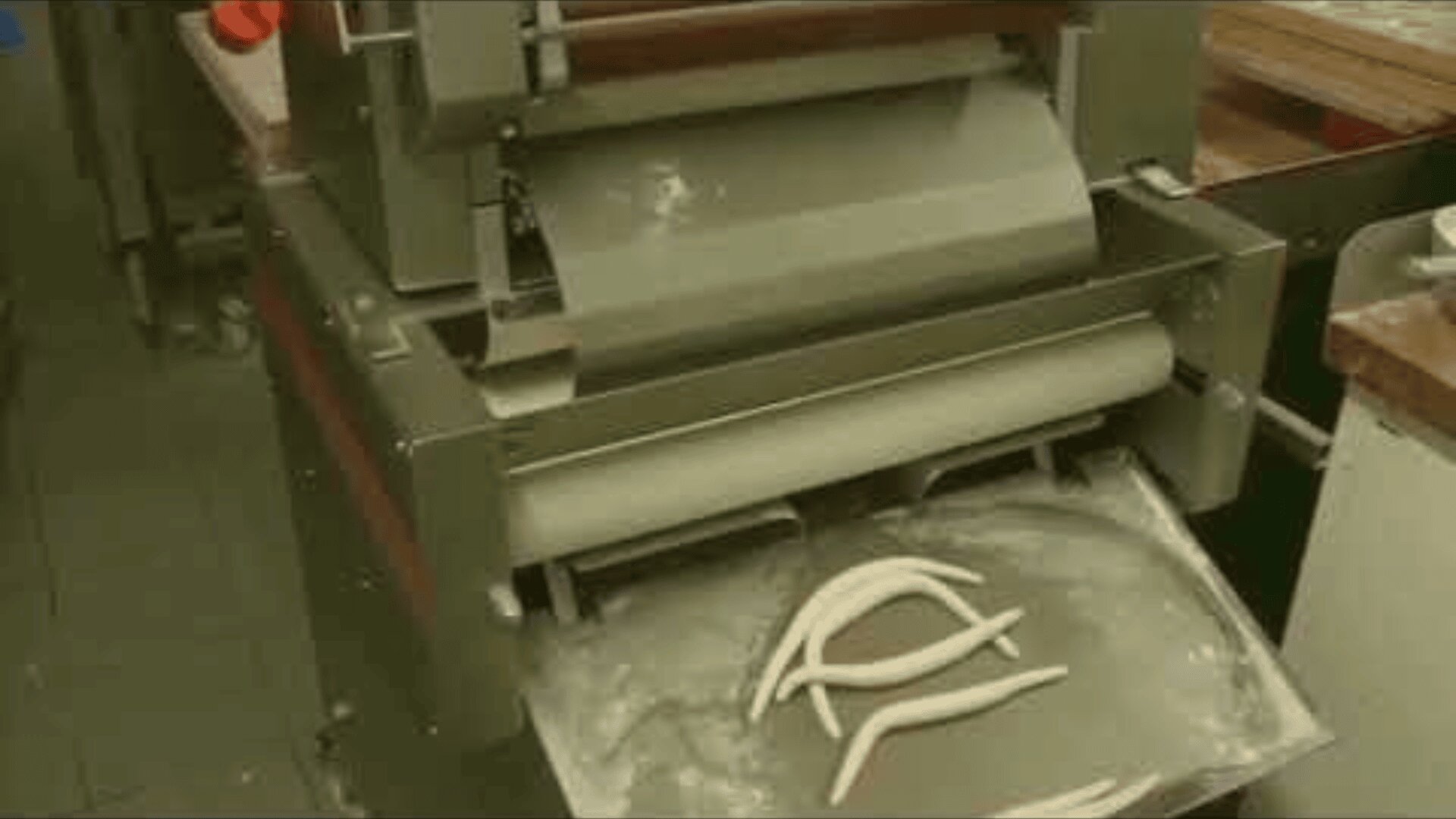 Eine handelsübliche Teigmaschine in einer Küche drückt Teigstücke auf ein darunter liegendes Tablett, auf dem Mehl verstreut ist.