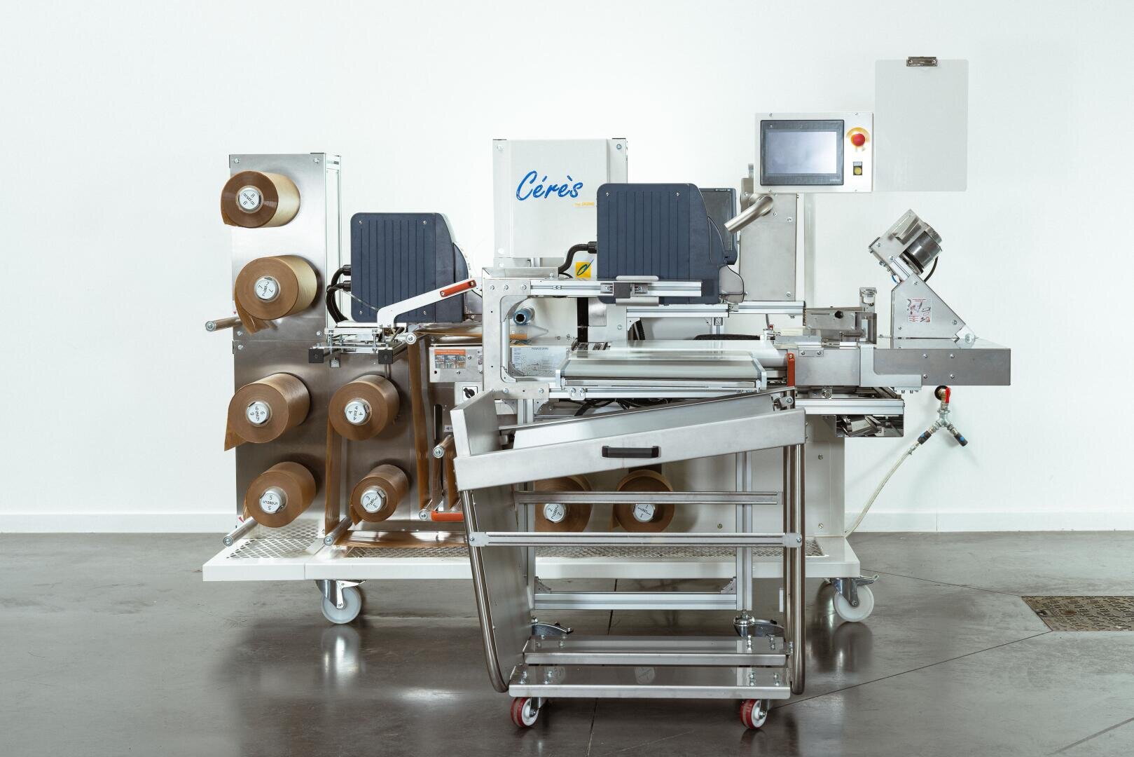 A stainless steel industrial packaging machine with multiple large rolls of brown paper, a touchscreen control panel, and various mechanical components, set on a polished concrete floor against a white wall.
