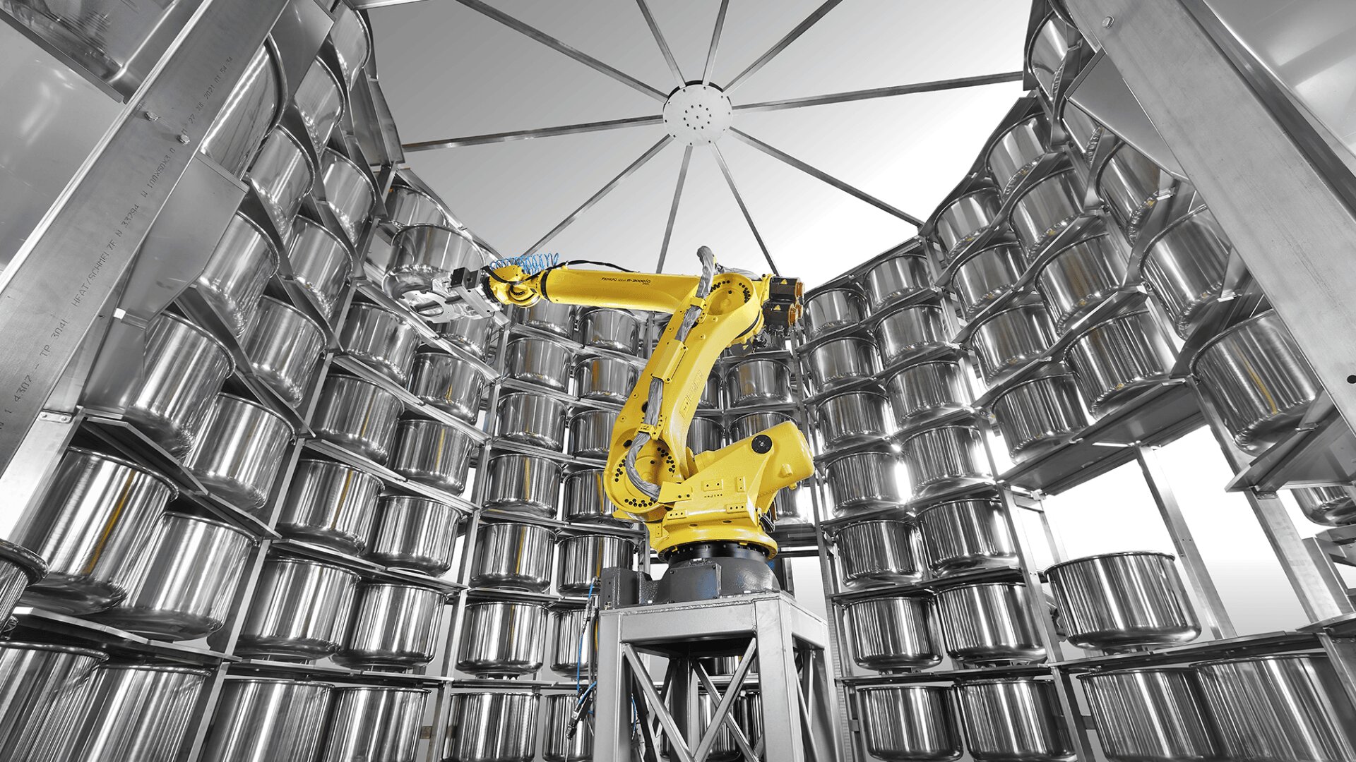 A yellow industrial robotic arm operates inside a circular, metal-walled chamber lined with rows of shiny, reflective containers stacked on shelves. Bright, even lighting highlights the clean, high-tech environment.