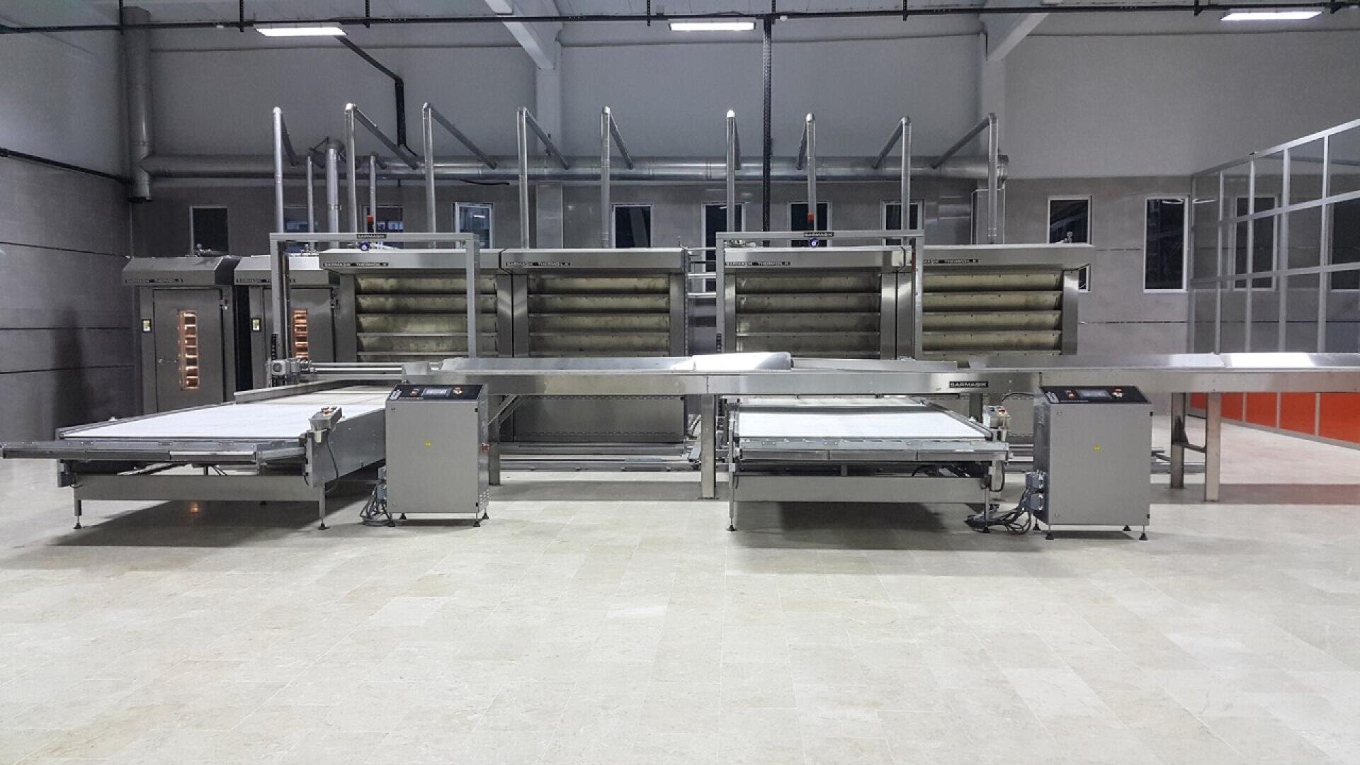 Large industrial bakery equipment with stainless steel conveyor belts and ovens in a spacious, modern facility with tiled floors and high ceilings.