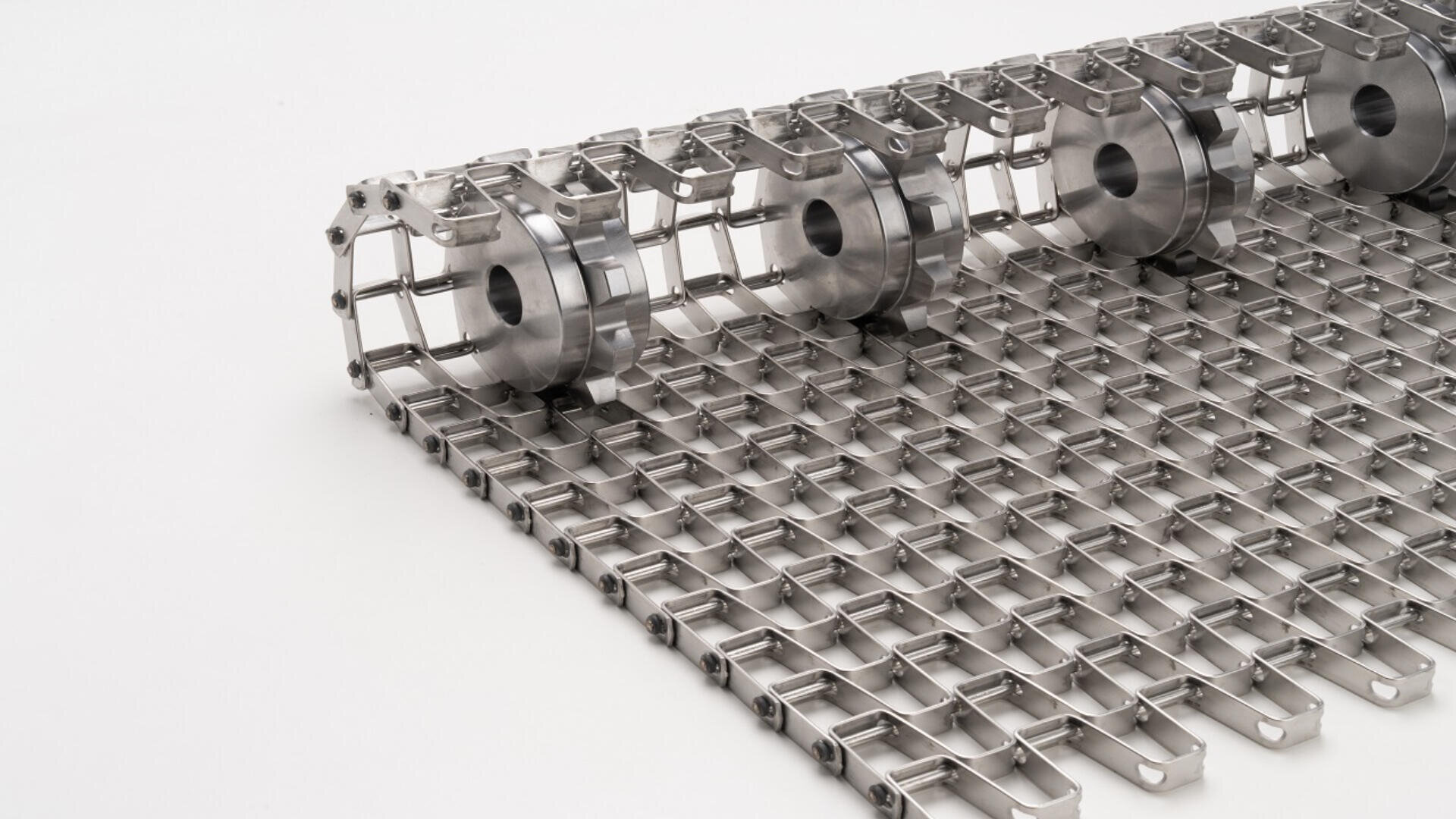 Close-up of a stainless steel conveyor belt with a mesh pattern and cylindrical rollers along one edge, set on a plain white background.