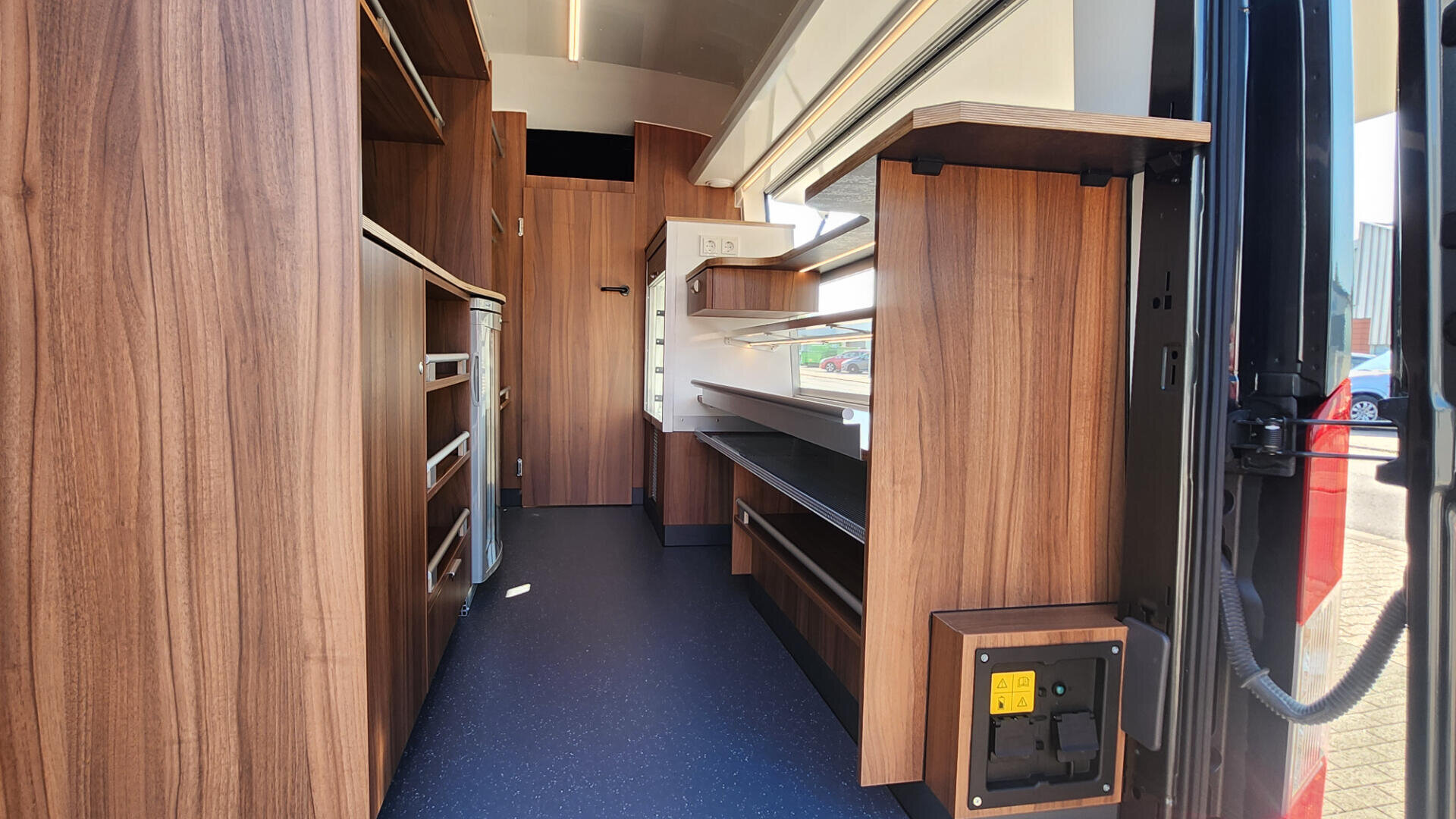 Innenraum eines mobilen Ladens oder Food Trucks mit holzgetäfelten Schränken und Regalen, dunkelblau gesprenkeltem Fußboden und großen Fenstern auf der rechten Seite, die natürliches Licht hereinlassen.