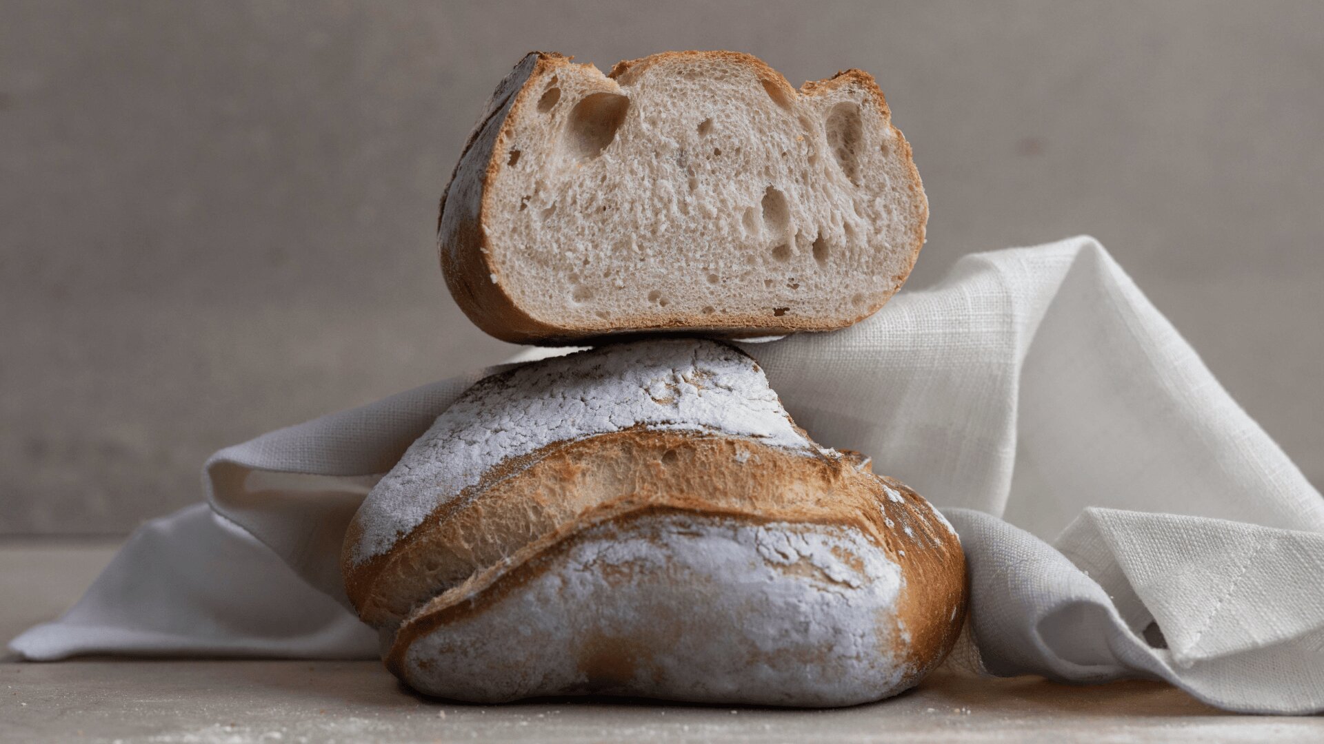 Ein rustikaler, mit Mehl bestäubter Laib Brot wird mit einem halbierten Stück belegt, dessen luftige Krume sichtbar ist. Beides liegt auf einer hellen Stoffserviette vor einem neutralen Hintergrund.