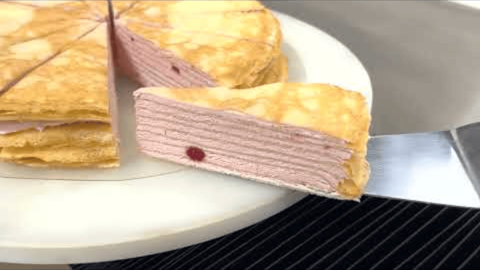 Ein Stück geschichteter Krepptorte mit hellrosa Füllung und einem einzelnen roten Fleck wird auf einem weißen Teller aus der ganzen Torte gehoben.