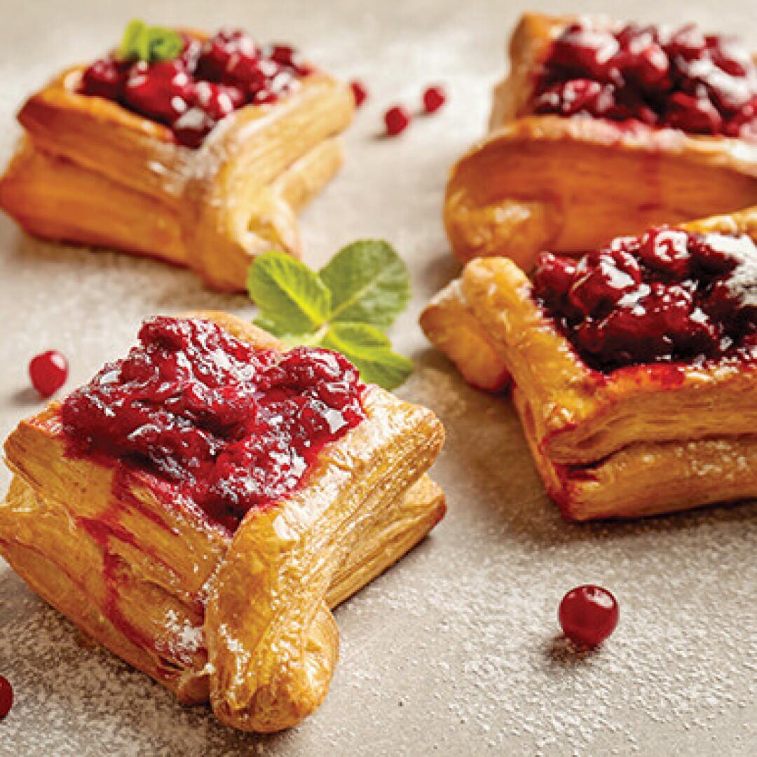 Four square puff pastry pockets with red berry compote are arranged on a lightly powdered surface, garnished with a sprig of mint and sprinkled with red berries.