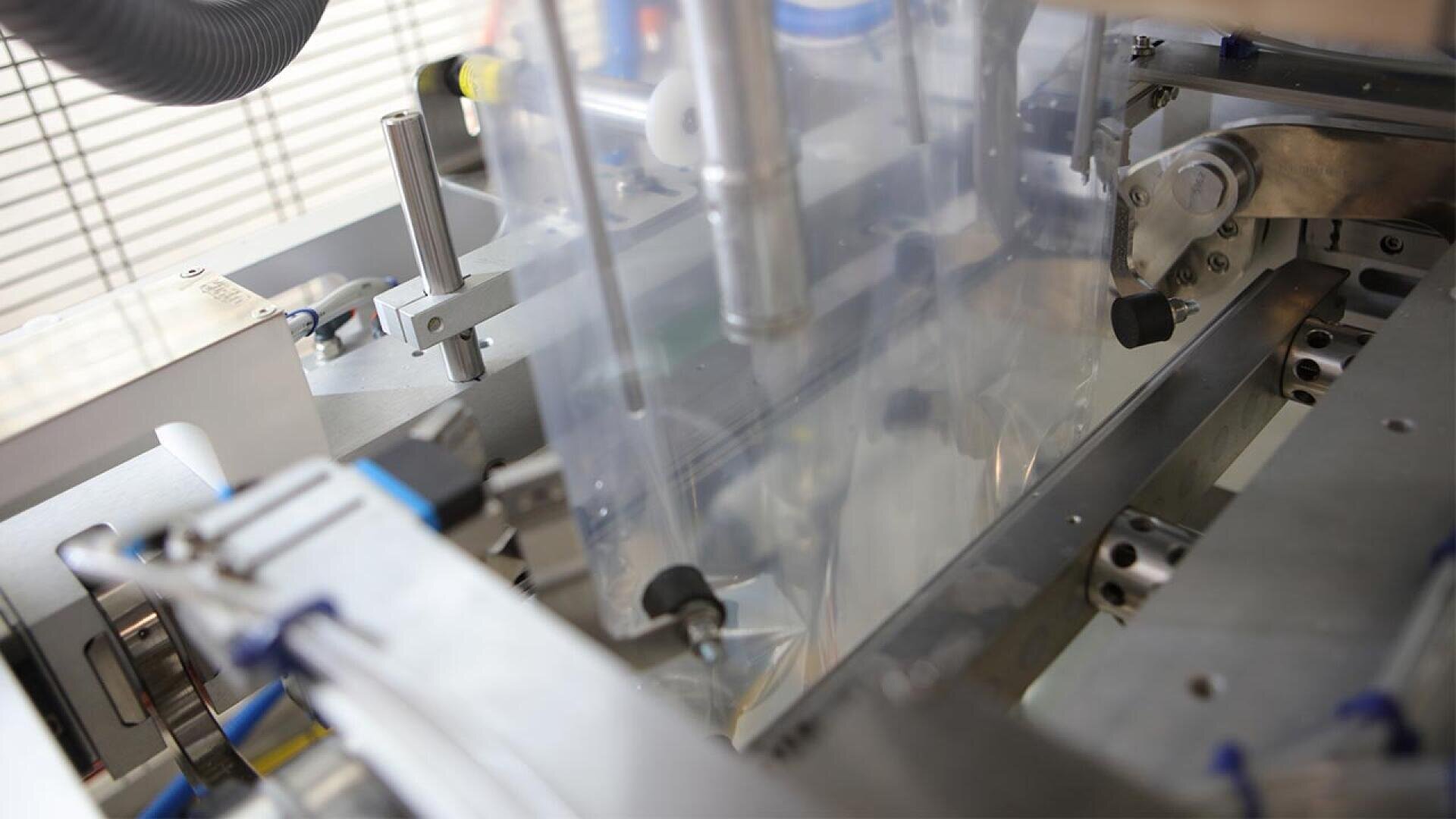 Close-up of an automated packaging machine with transparent film being sealed and advanced by metal rollers, showing various mechanical parts and industrial components.