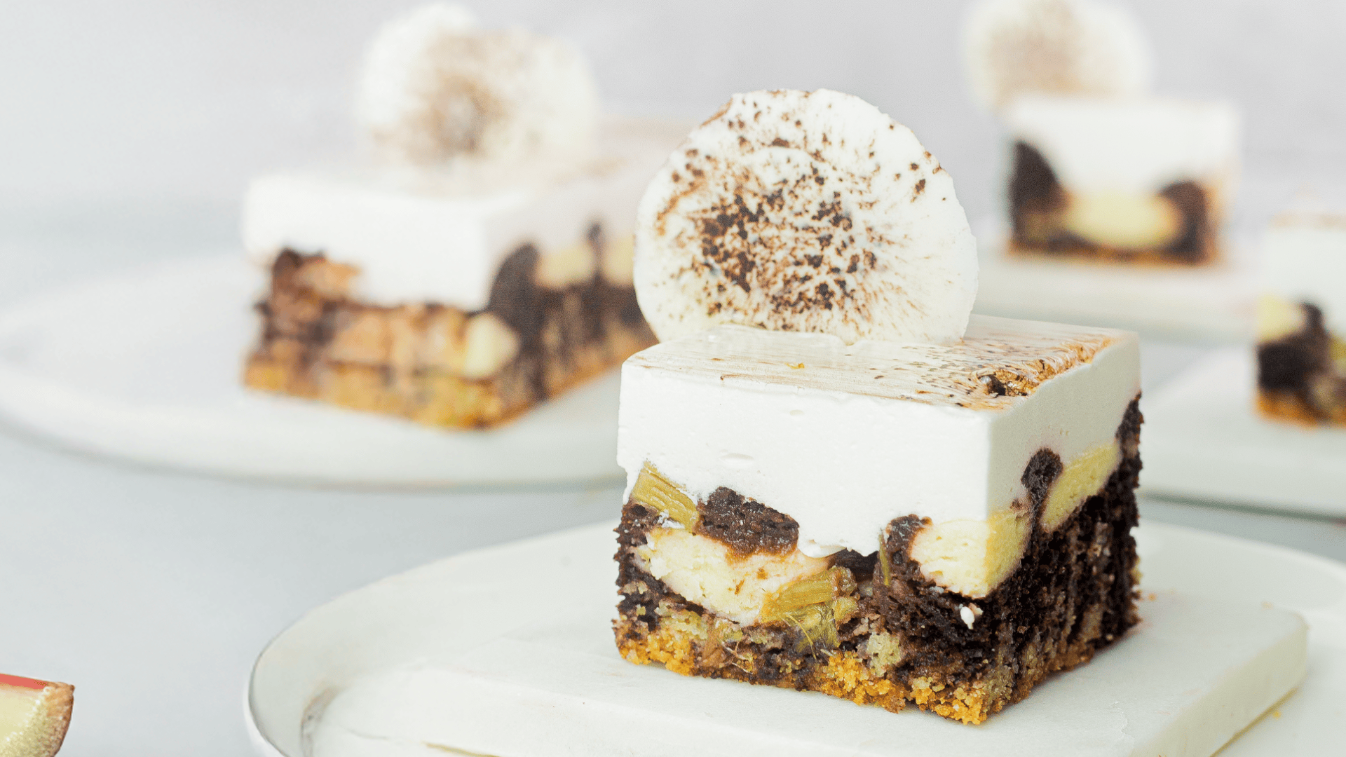 A close-up of a square dessert with layers of cake, fruit, chocolate, and thick white frosting, topped with a round chocolate-dusted decoration, on a white plate with similar desserts in the background.