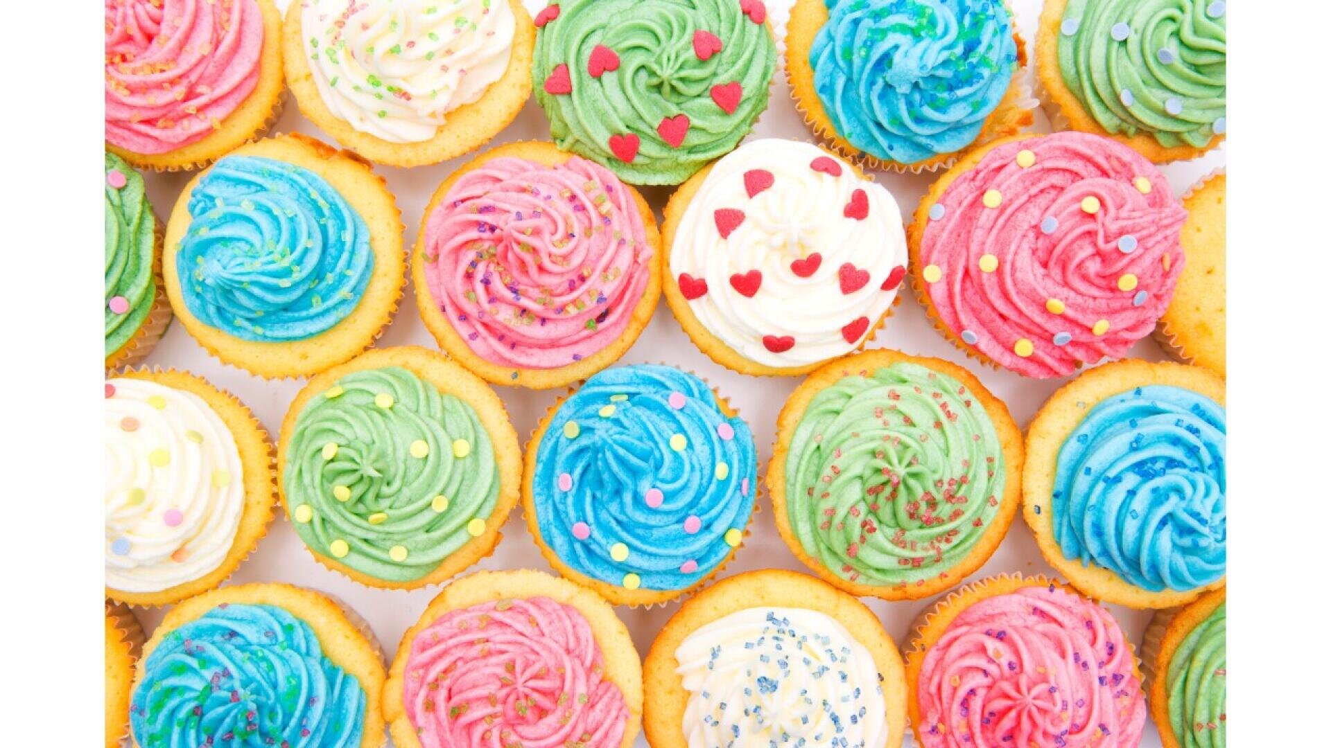 A top view of colorful cupcakes with swirled frosting in pink, blue, green, and white, decorated with sprinkles and candy hearts, arranged closely together.