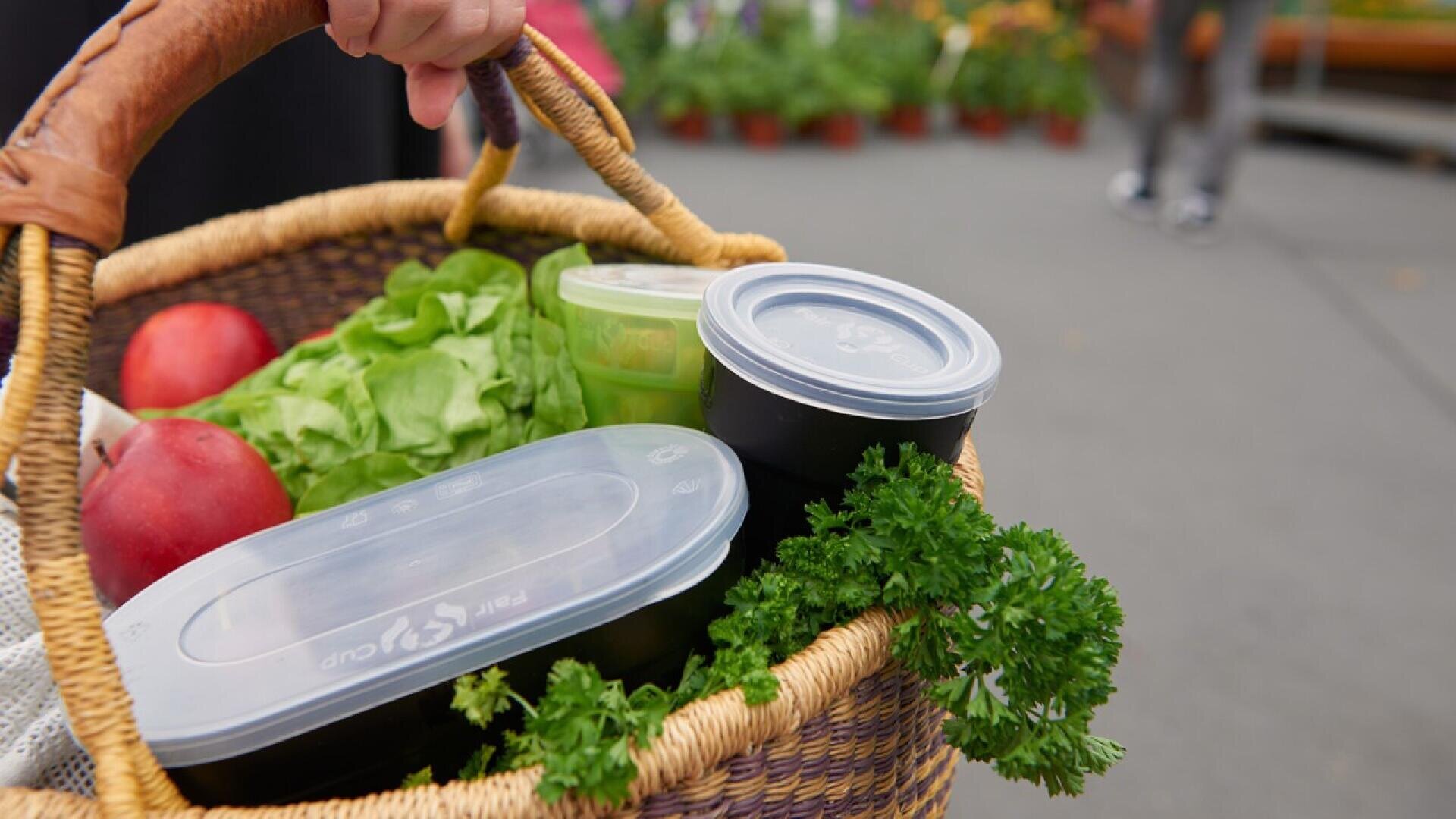A close-up of a woven basket filled with leafy greens, parsley, two red apples, and several plastic food containers with lids. A hand holds the basket, and the background shows an outdoor market setting.