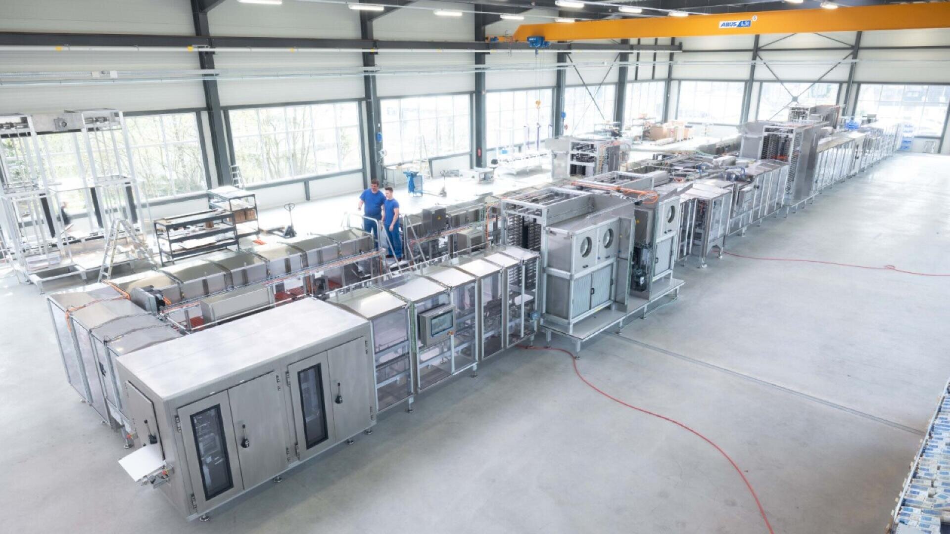 A large industrial facility with stainless steel machinery and equipment arranged in rows; two workers in blue uniforms stand near the machines, overseeing or assembling components in a bright, spacious room.