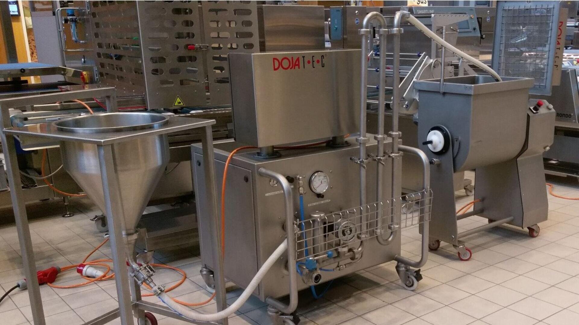 A stainless steel industrial food processing machine with pipes, gauges, a funnel, and control panels is set up in a tiled facility, with orange cables and metal equipment surrounding it.