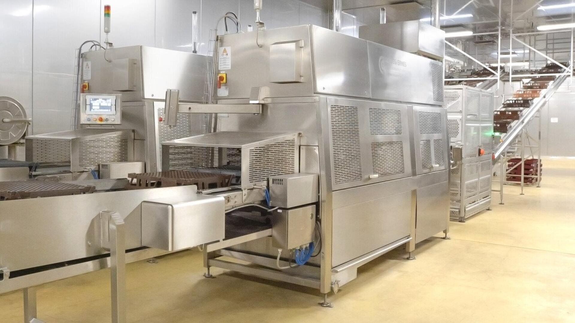 Large industrial food processing machines made of stainless steel are set up in a clean, well-lit factory with yellow flooring and metal stairs leading up to an elevated platform in the background.