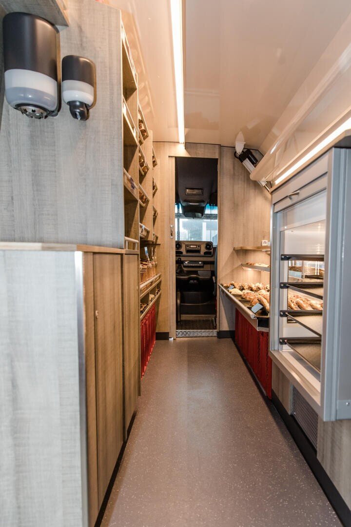 Ein schmaler Innenraum eines modernen Imbisswagens mit Holzregalen auf der linken Seite, einer Glasvitrine mit Gebäck auf der rechten Seite und einem Blick in die Fahrerkabine am anderen Ende.