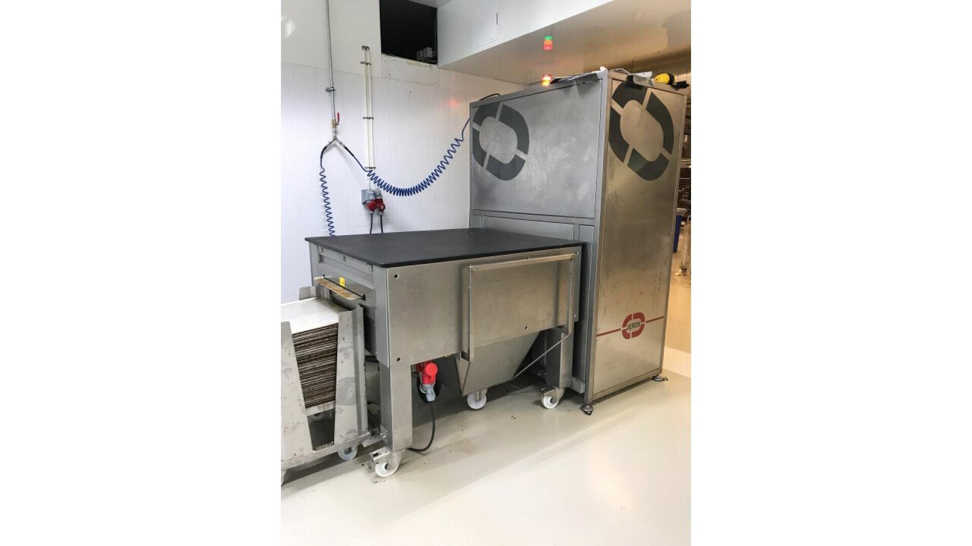 A stainless steel industrial machine with wheels and a black tabletop stands next to a tall enclosed unit with circular symbols. A coiled blue hose is mounted on a white wall nearby.