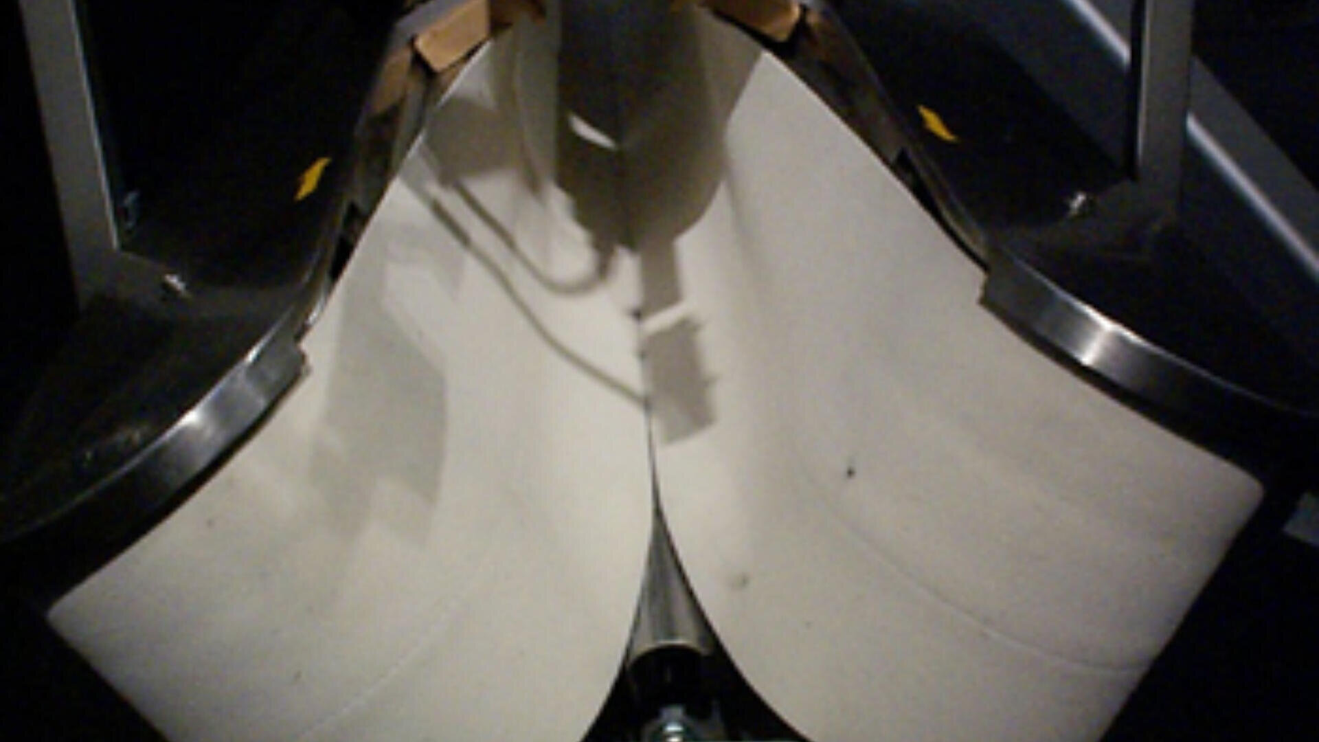 Nahaufnahme von zwei großen, weißen Förderbändern, die sich in einem nach unten gerichteten Winkel treffen, eingerahmt von Metallkanten, möglicherweise Teil einer industriellen Verarbeitungs- oder Sortiermaschine.