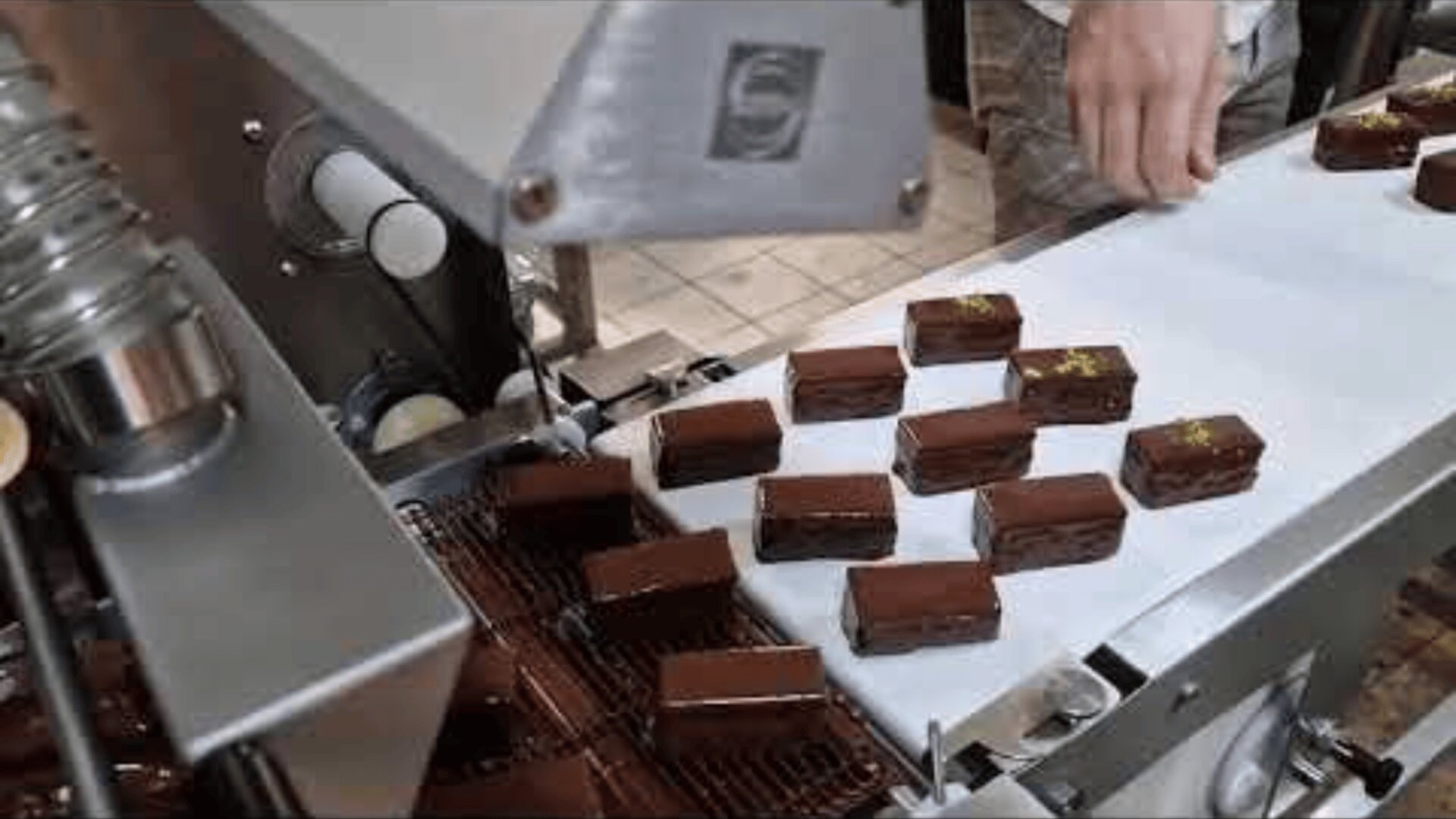 Chocolate-covered pastries moving along a conveyor belt in a factory, with a persons hand visible arranging the pastries as they are coated and sorted.