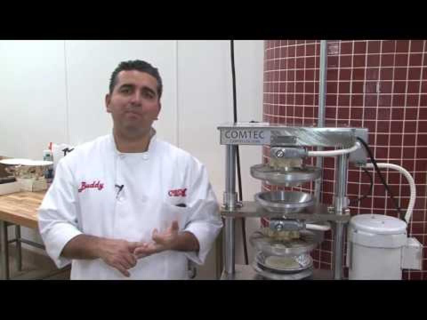 Ein Koch im weißen Kittel steht neben einer kommerziellen Teigpresse in einer Küche mit weißen Wänden und einem rot gefliesten Bereich.