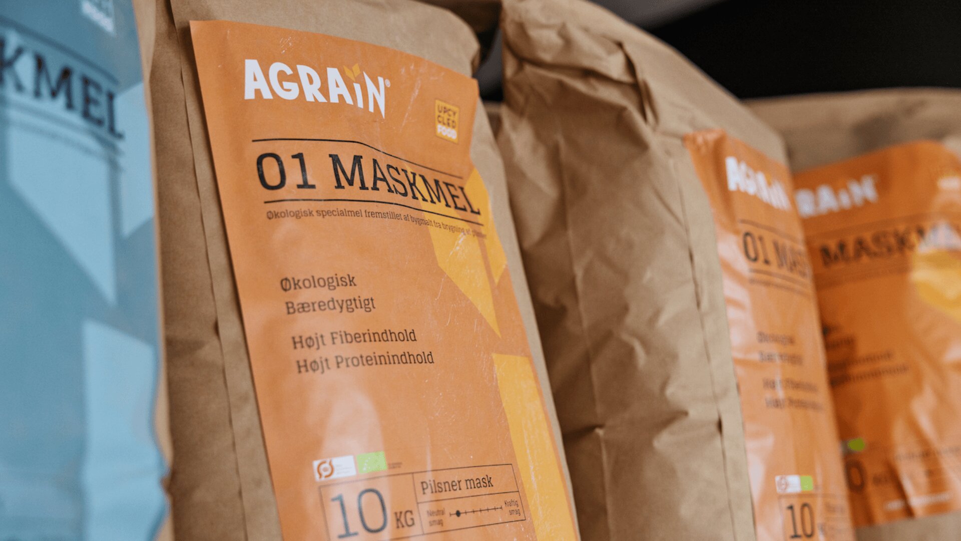 Brown paper bags of AGRAN 01 Maskmel, an organic and high-protein flour product, are displayed in a row. The packaging includes text in Danish and features orange and yellow labels.