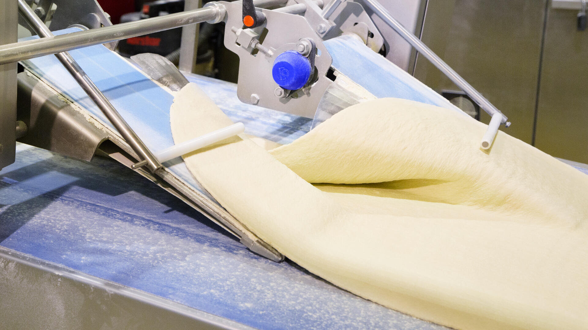Sheet of dough being processed and flattened by an industrial machine in a food production facility, with flour dust visible on the equipment.